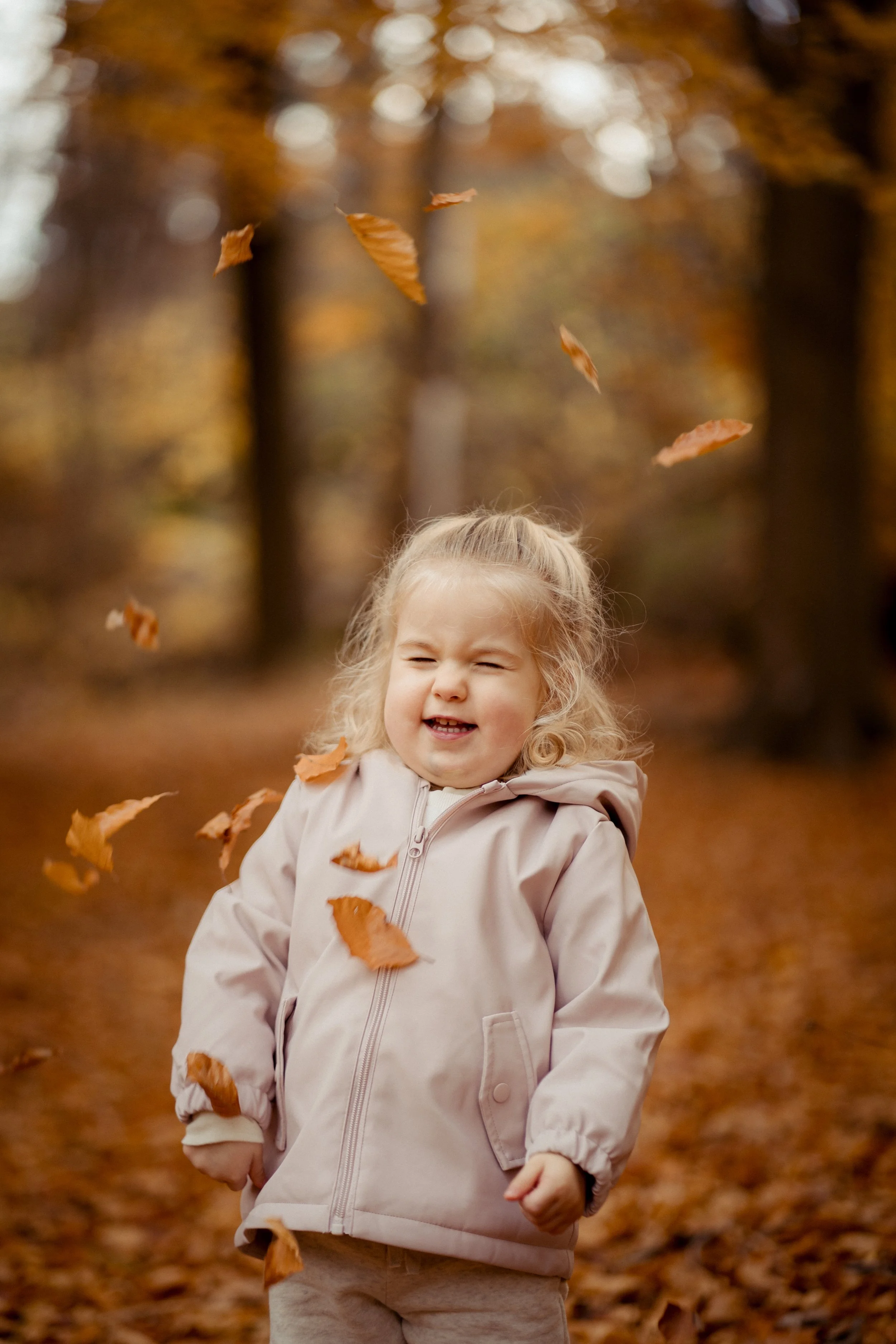 A young girl with curly blonde hair, wearing a beige jacket, smiling with eyes closed while playing with falling autumn leaves in a wooded park during fall.
