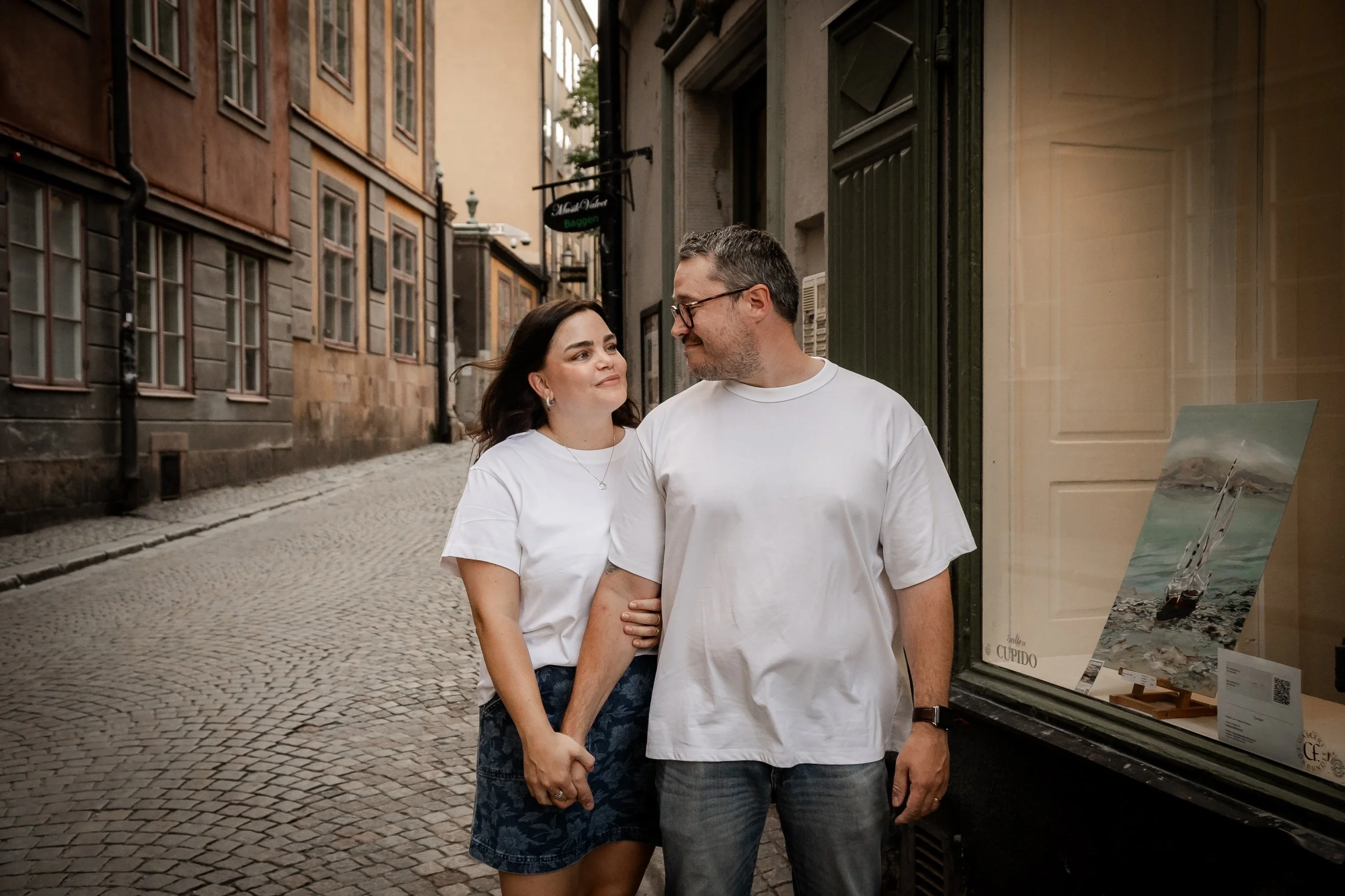 A couple holding hands on a cobblestone street at sunset, smiling and looking at each other, with old buildings in the background.