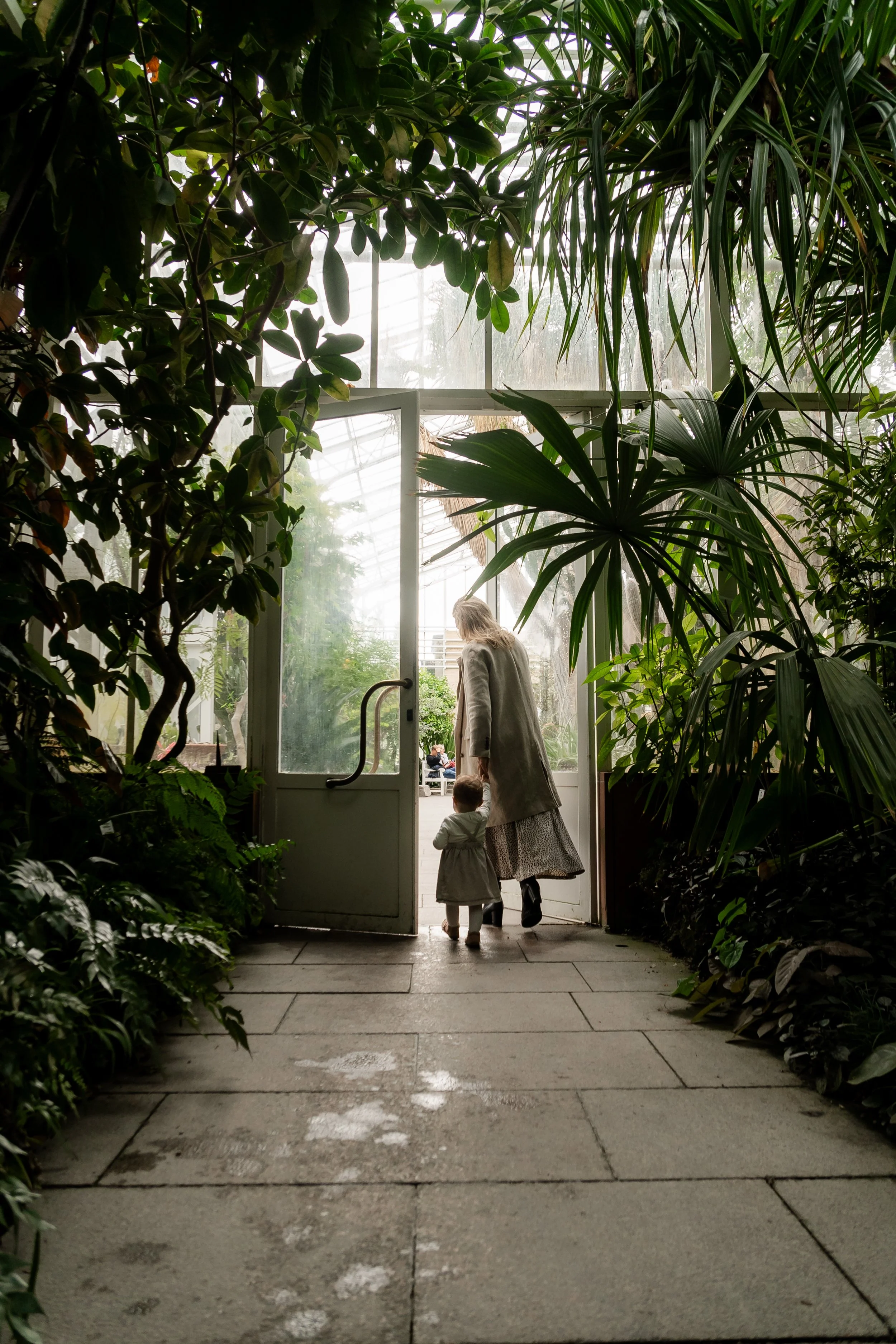 A woman and a child walking through a glass door into a lush indoor garden or greenhouse filled with various green tropical plants.
