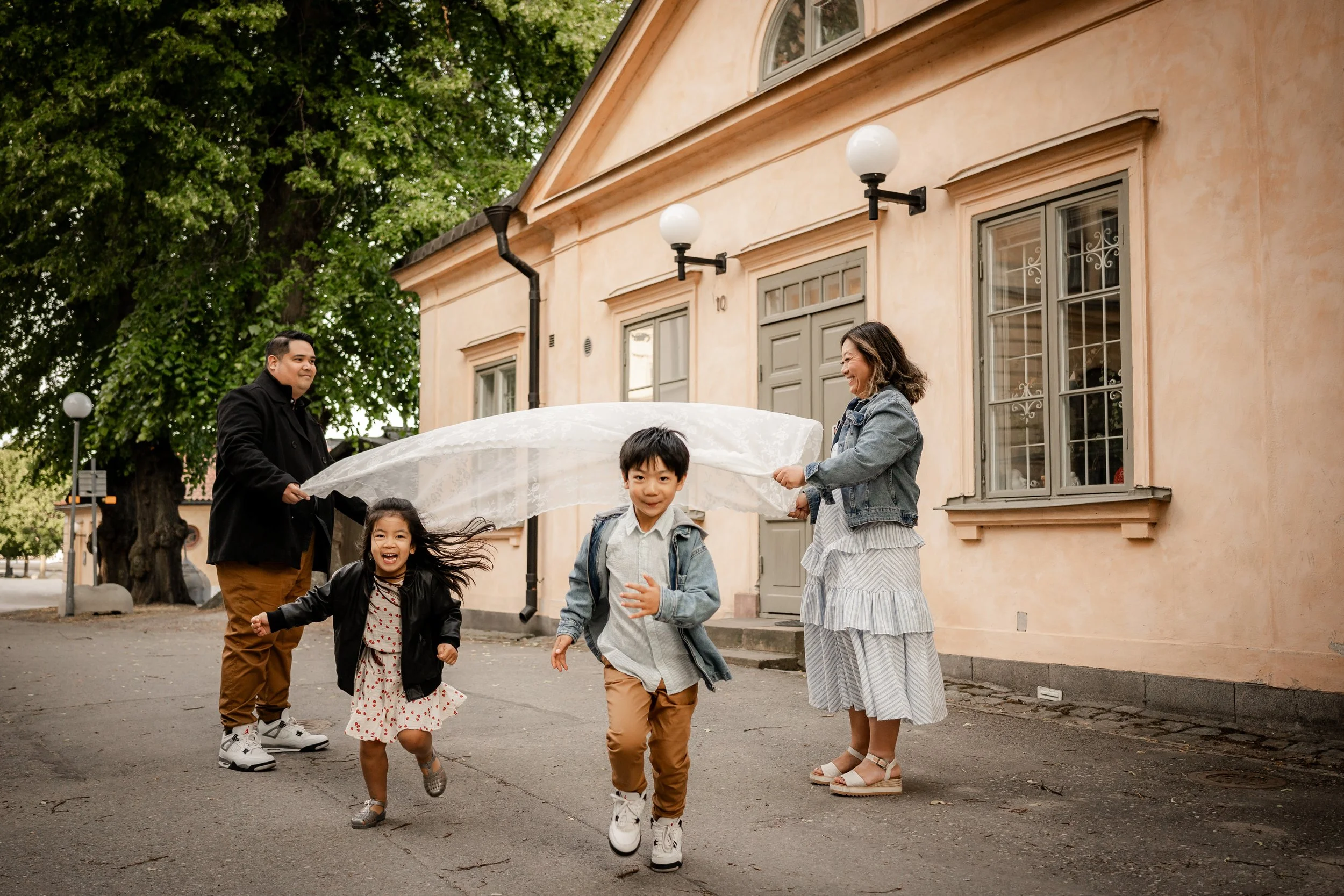 A family of four enjoying time outdoors, running and playing with a white cloth or sheet, in front of a beige house with large windows and decorative black lamp posts. The children are smiling and appear happy.