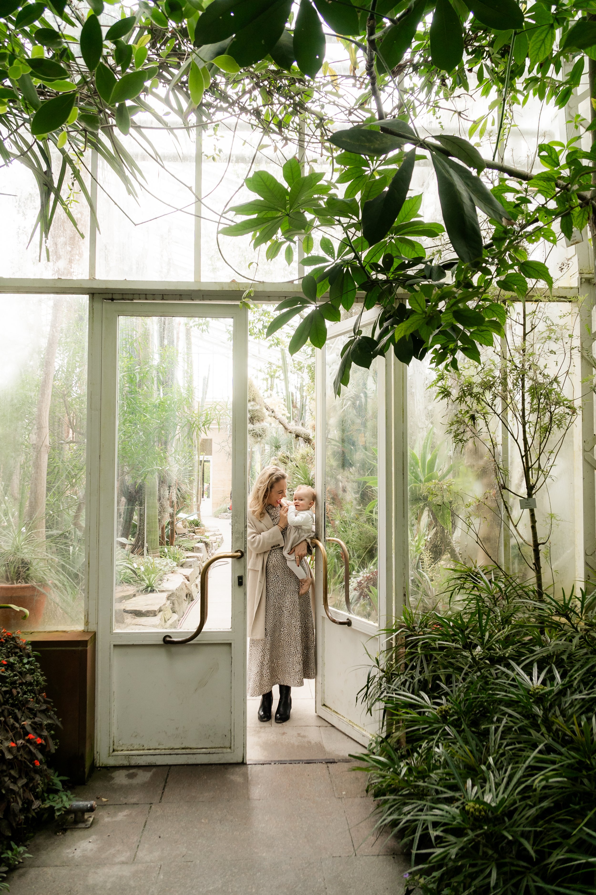 A woman holding a child at the entrance of a glass greenhouse filled with lush green plants and trees.