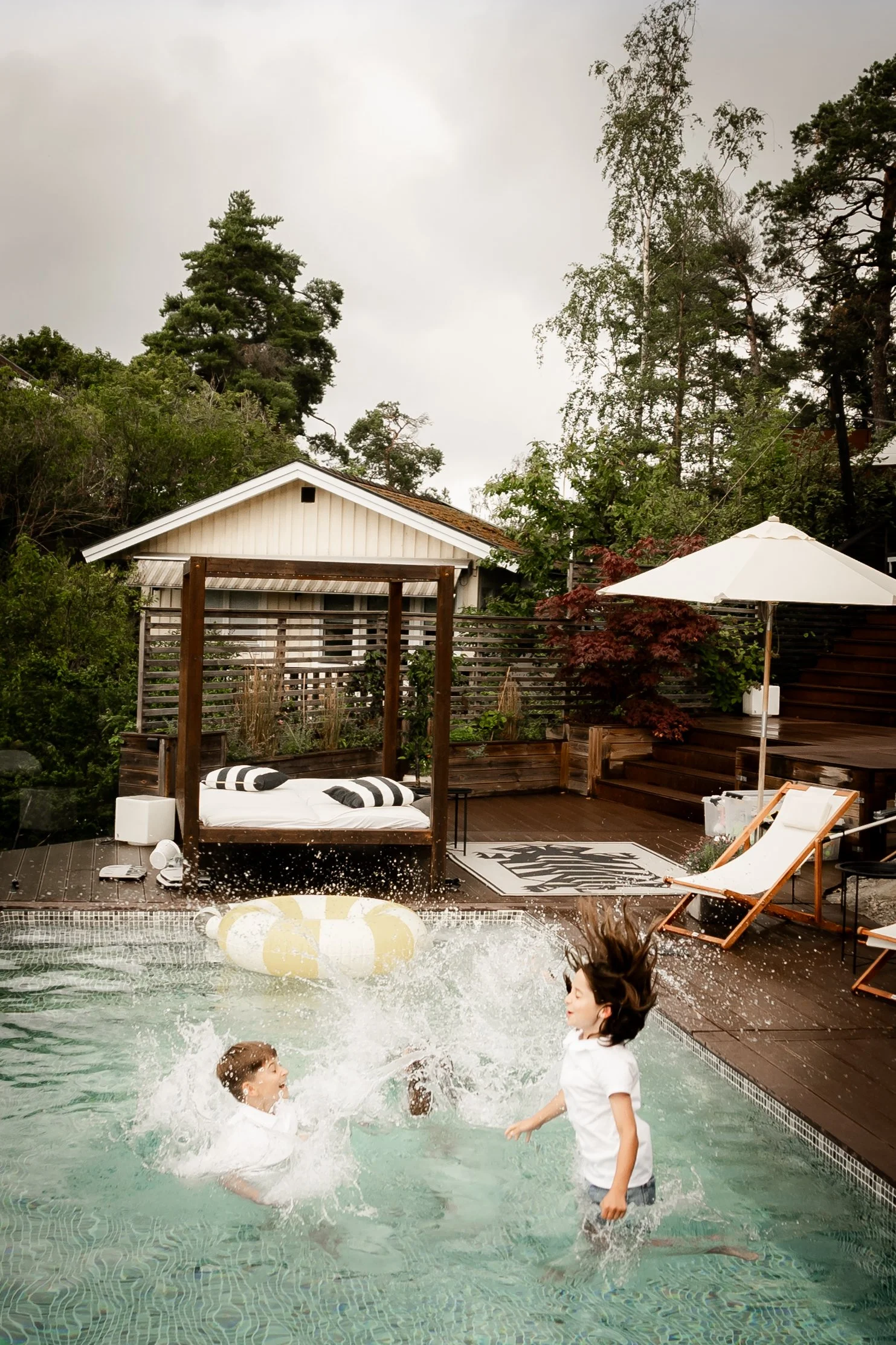 Two children playing in a backyard swimming pool with splashing water. The backyard has outdoor furniture, a large white umbrella, and lush greenery with trees and shrubs surrounding the area.