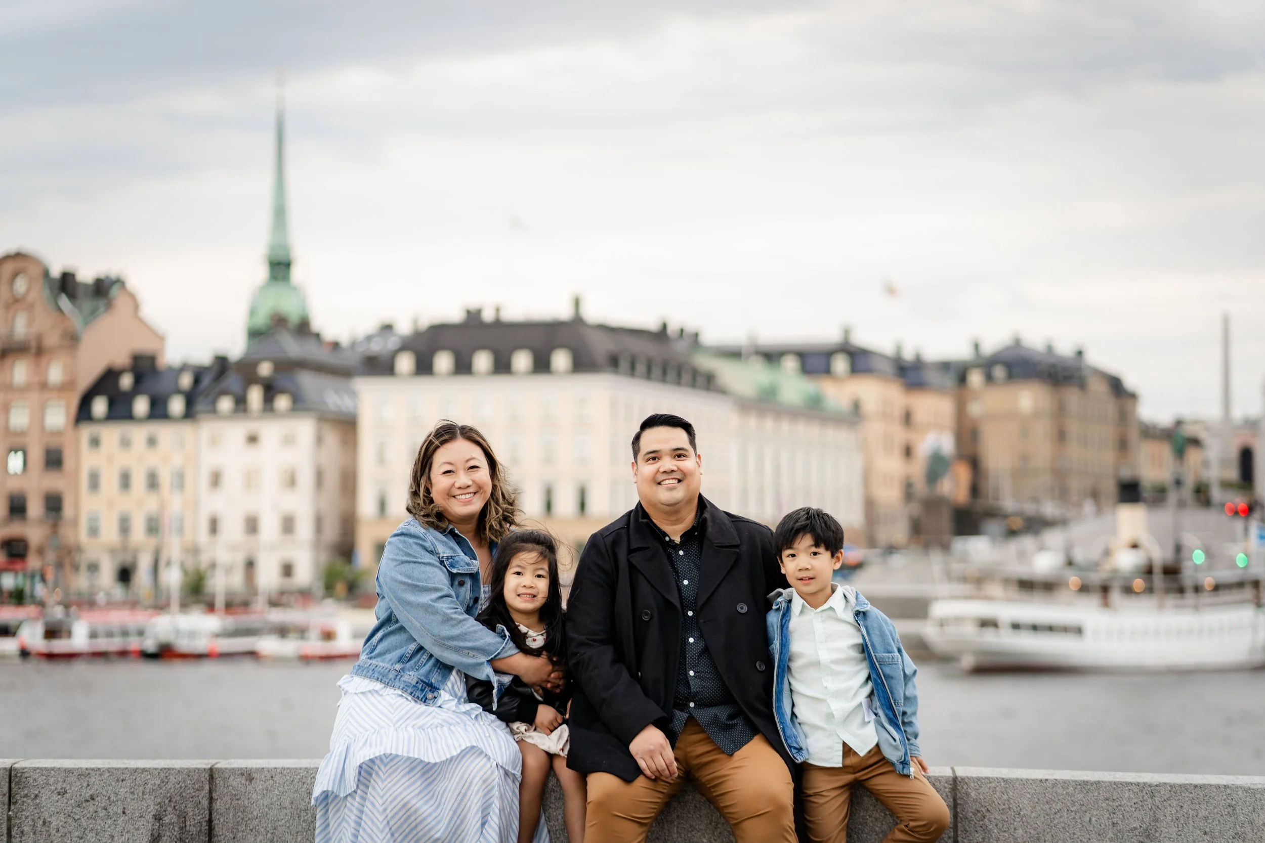 Family of four sitting on a stone wall by a river with city buildings and a church steeple in the background.