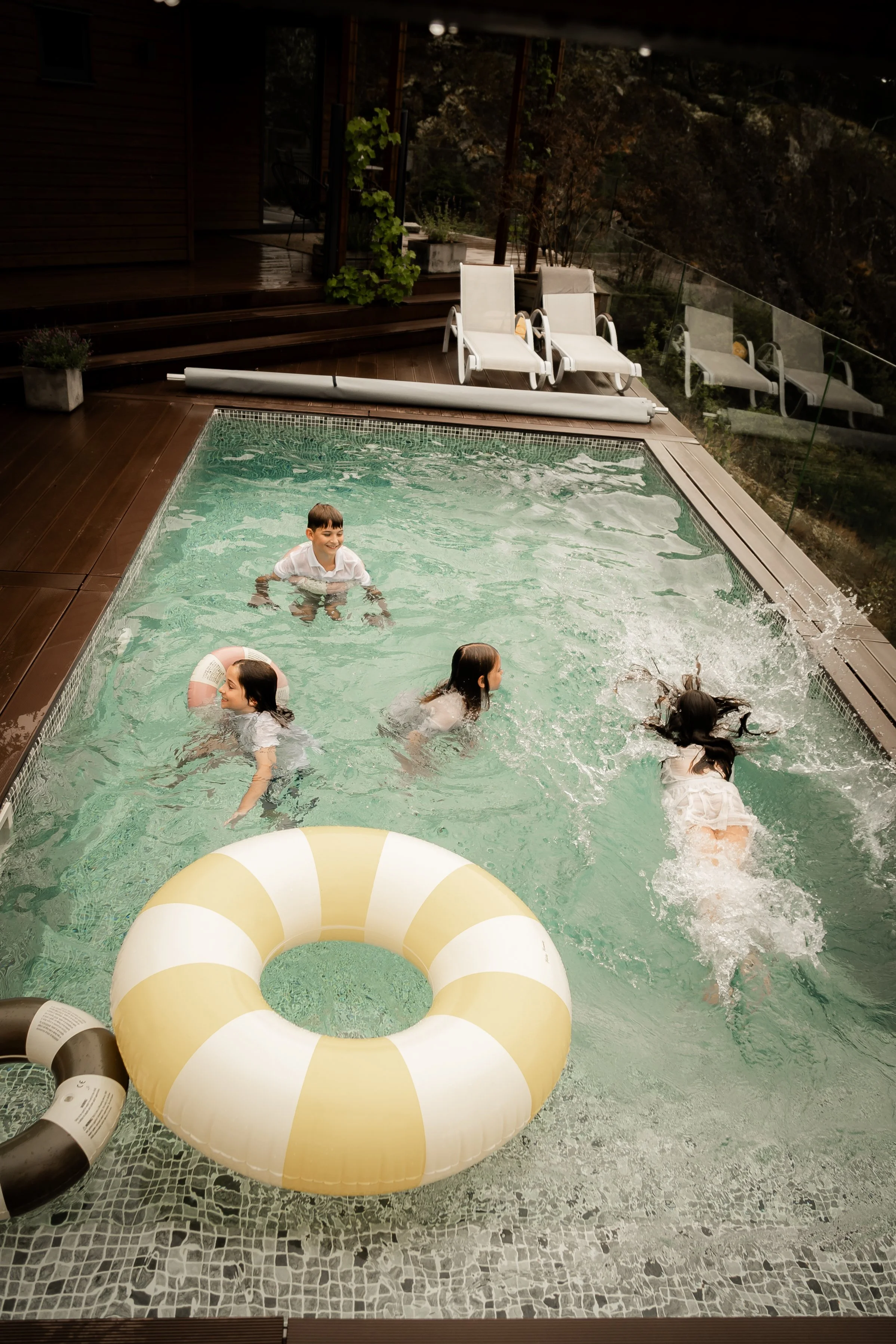 Children playing and swimming in a backyard pool surrounded by a wooden deck, poolside lounge chairs, and patio plants, with a glass railing overlooking trees.