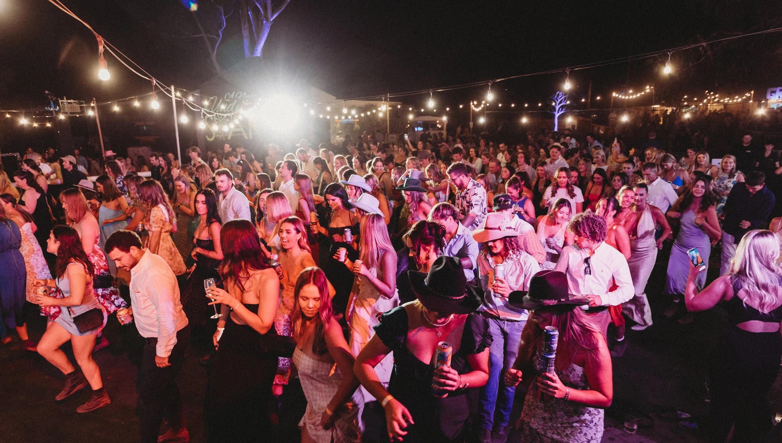 Crowd of people dancing and socializing at a nighttime outdoor party with string lights and a bright light in the background.