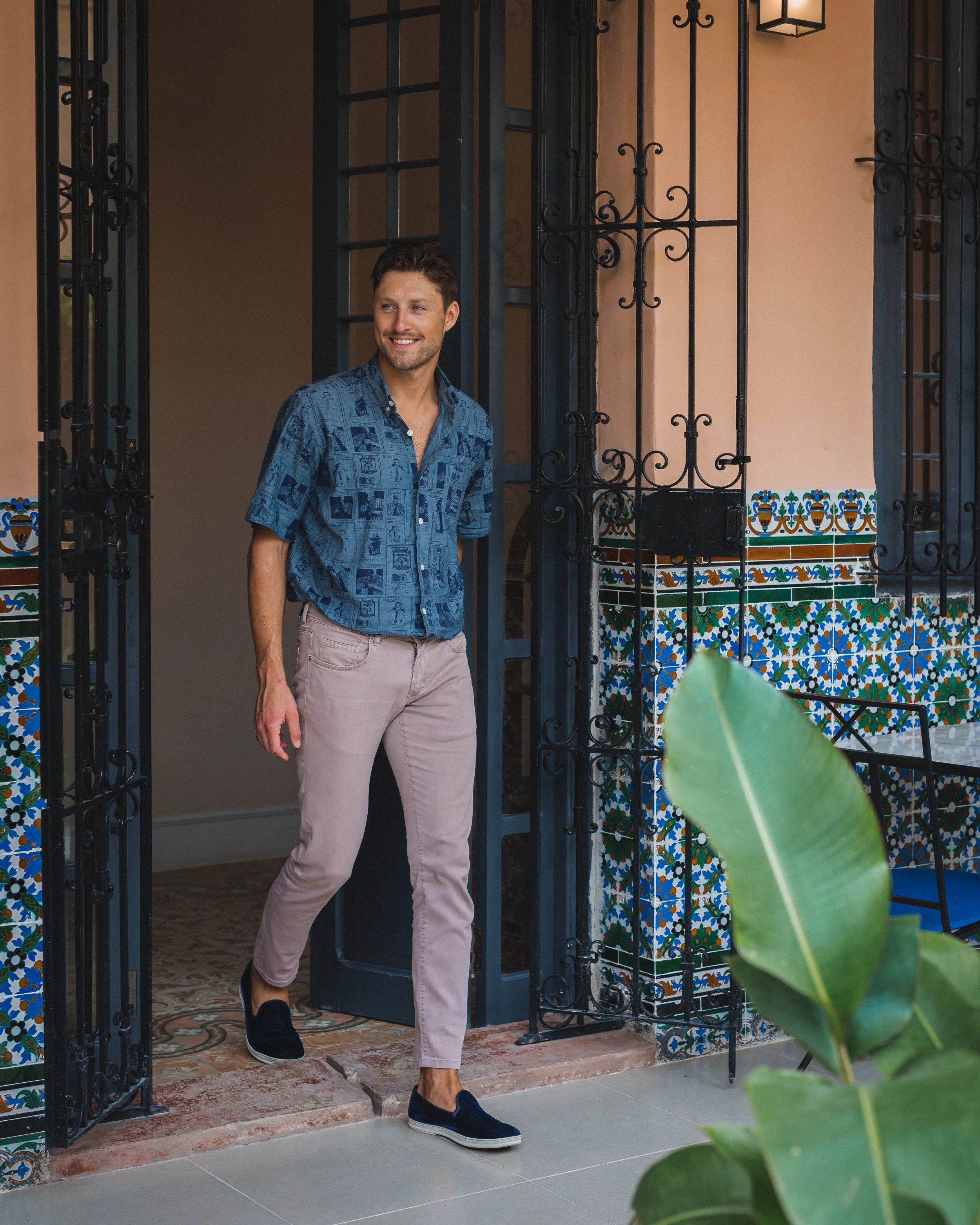 Un hombre con camisa azul de manga corta estampada y pantalones claros sale sonriente de una casa con portón de hierro negro y decoración de azulejos de colores.