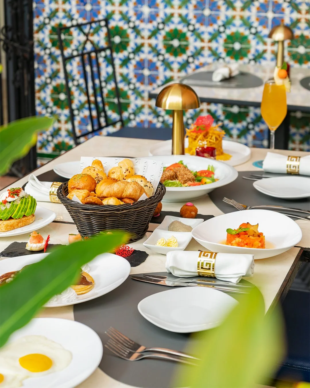 Una mesa de comedor elegantemente puesta con varios platos, incluyendo una cesta de pan, ensaladas, pasta, huevos y bebidas, en un restaurante con una colorida pared de azulejos estampados.