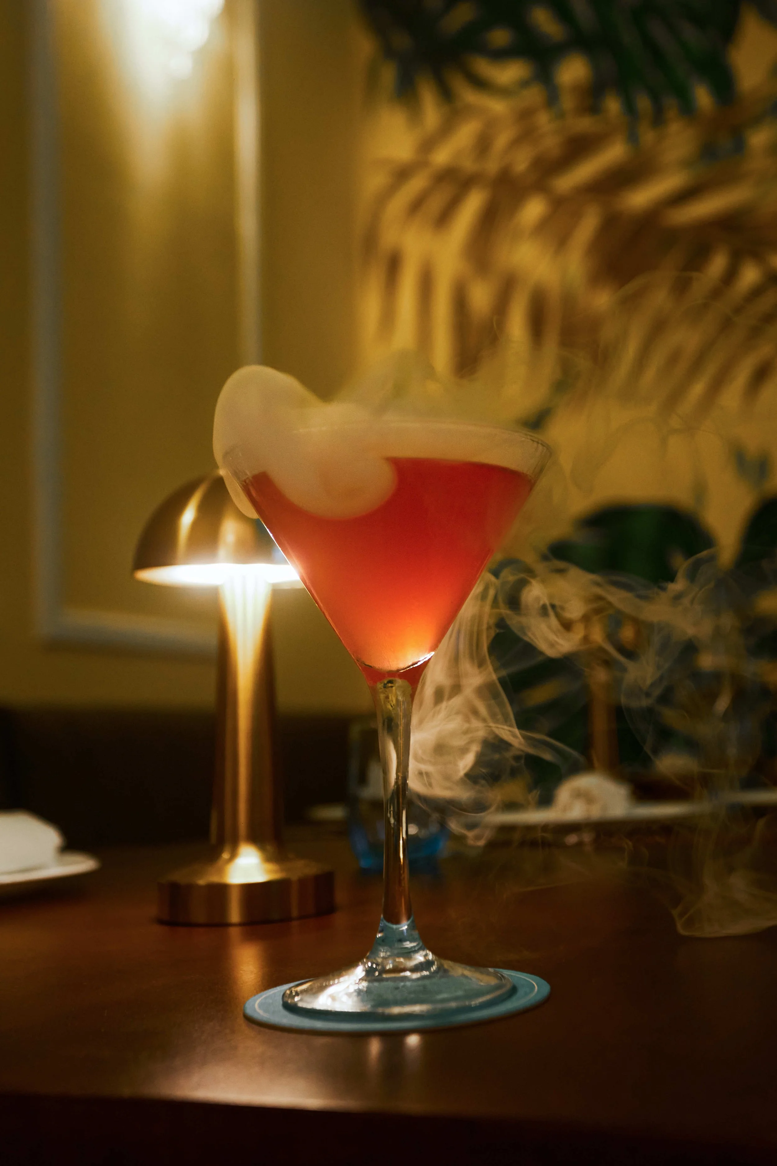 Cóctel en copa de martini con líquido rojo cereza, coronado con vapor o humo blanco, sobre una barra o mesa con una lámpara dorada y una planta decorativa al fondo.