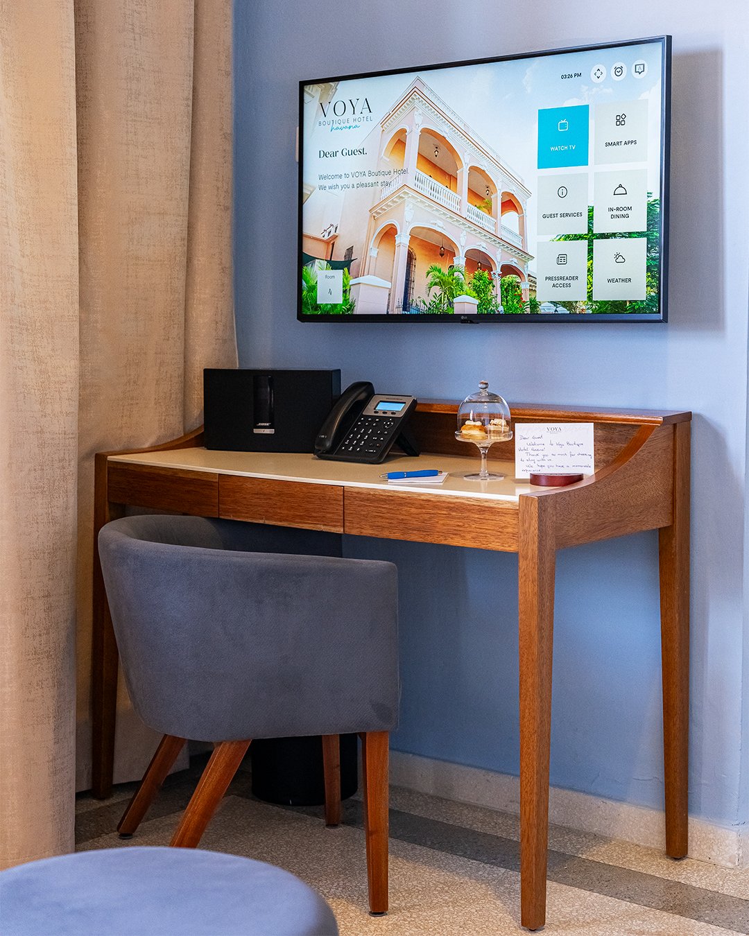 Hotel room workspace with a wooden desk, gray upholstered chair, a landline phone, a small black speaker, a glass dome with pastries, a blue pen, and a handwritten note, positioned beneath a mounted flat-screen TV displaying a hotel welcome message a