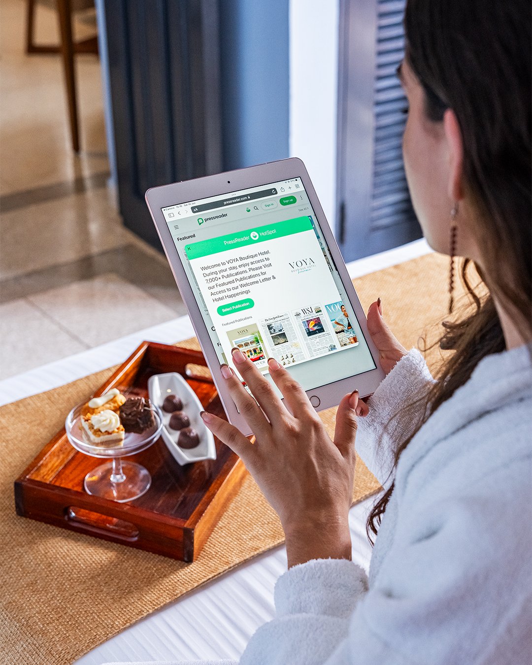 A woman in a white robe is using a tablet at a hotel bed with a tray of desserts nearby, including a glass of whipped cream topped dessert and chocolate truffles.