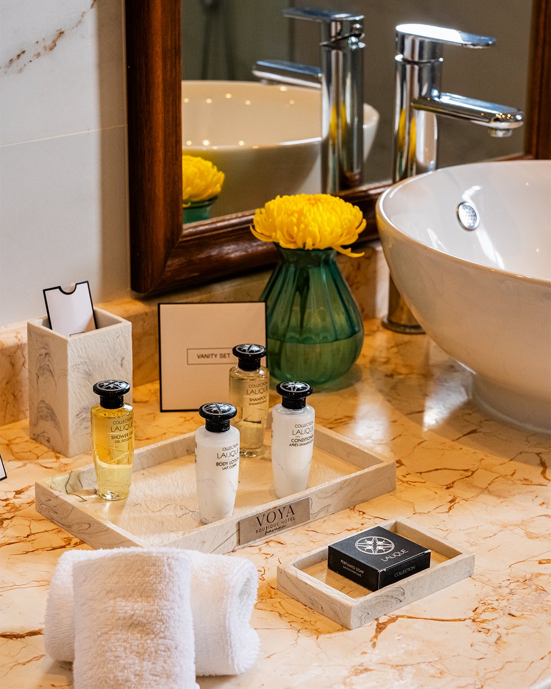 Bathroom countertop with yellow flowers in a green vase, a white vessel sink with a chrome faucet, small bottles of toiletries labeled Lalique, a mirror, a tissue box, a black box labeled Lalique, and a rolled white towel.