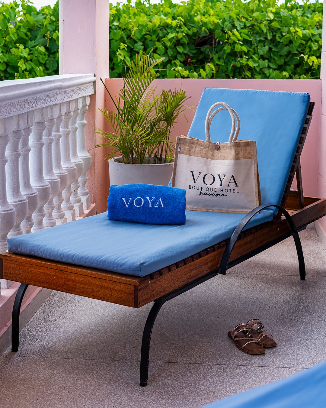 A wooden lounge chair with a blue cushion, placed on a balcony with pink walls. On the lounge chair, there is a beige tote bag with black text, a blue towel with white text, and a pair of brown sandals on the floor nearby. There is also a potted gree