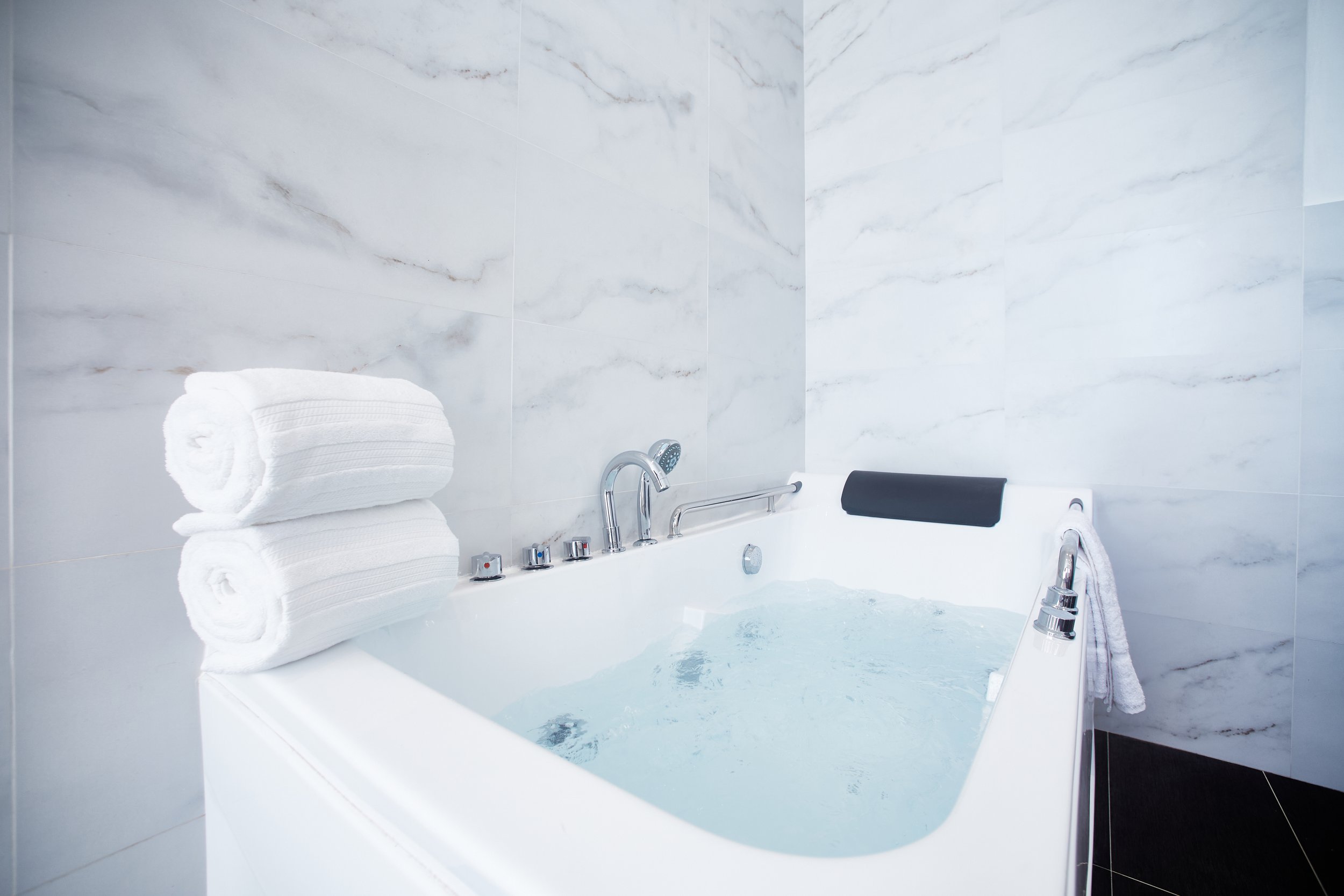 Luxury bathtub filled with water, with three rolled white towels stacked on the edge, a black headrest, and a white towel hanging on the side, in a bathroom with marble tile walls.