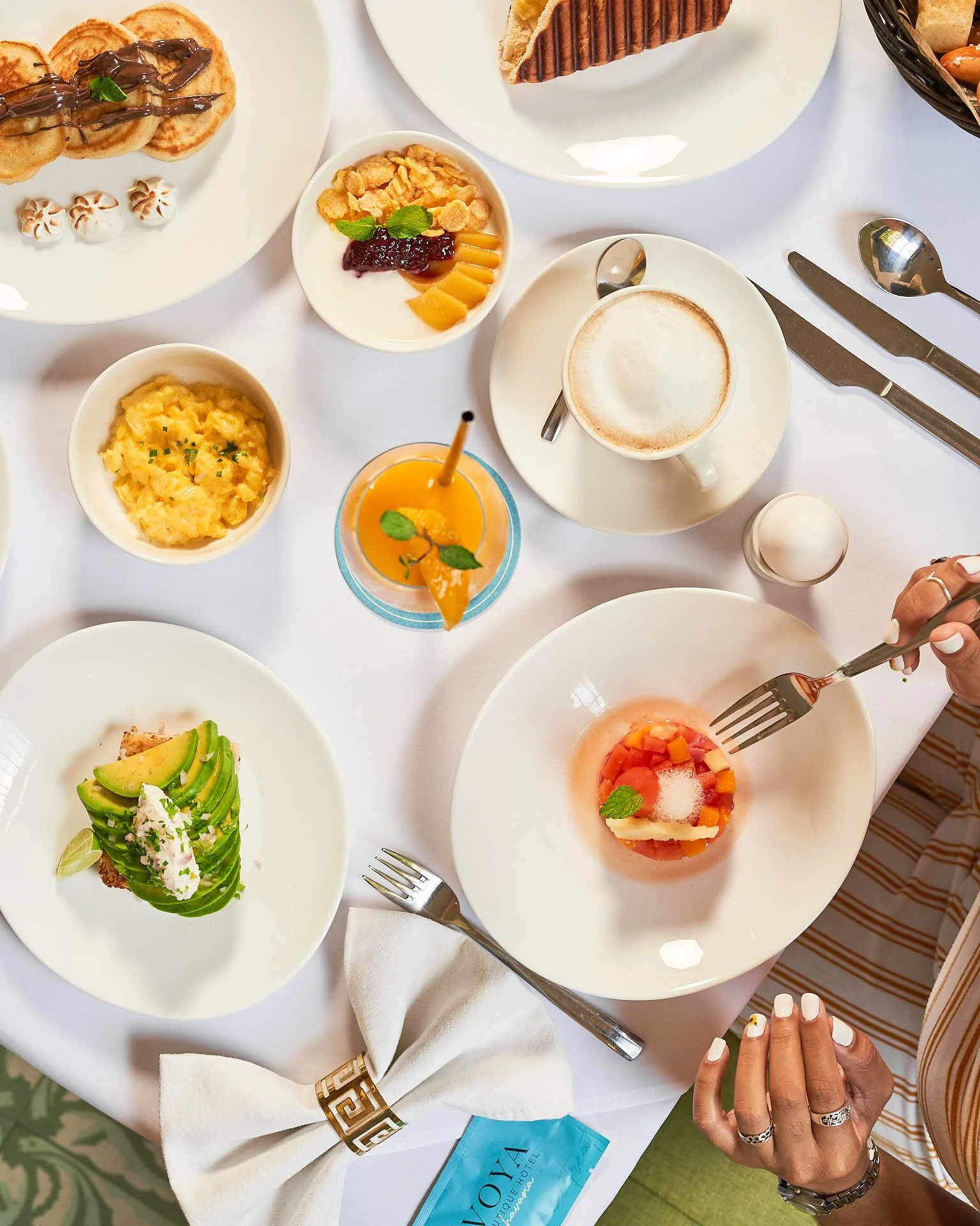 Una mesa con varios platos de desayuno, incluidos gofres con llovizna de chocolate, un bol de cereales con fruta y yogur, huevos revueltos, una tostada de aguacate, una ensalada de frutas, un vaso de zumo de naranja, un capuchino y un bol de tarta de tomate y verduras. En la mesa también hay cubiertos y una servilleta.