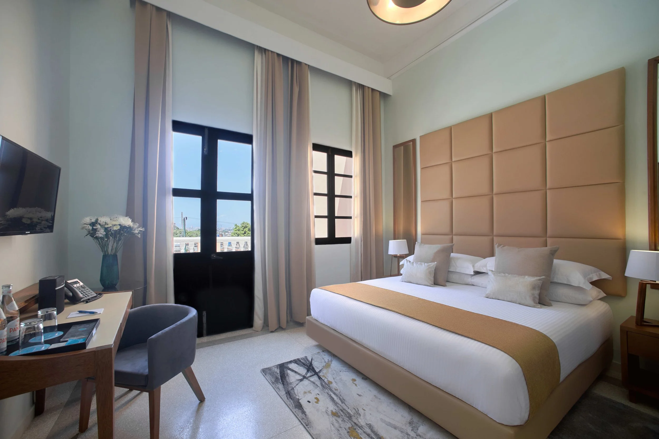 Hotel room with a large bed, beige headboard, white linens, multiple pillows, side tables with lamps, a window with white curtains, a desk with a chair, a TV, and a vase with white flowers.