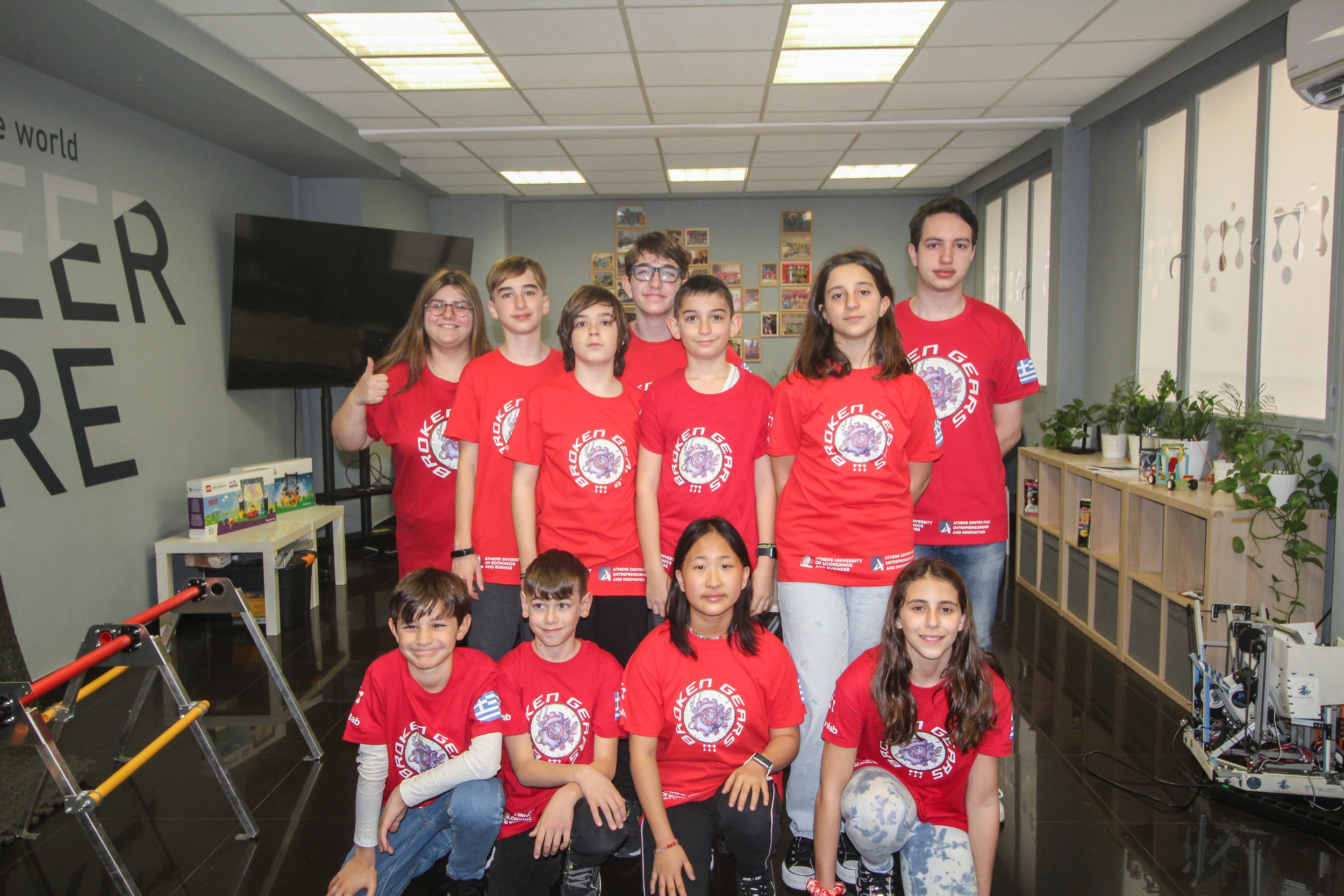 A group of children and teenagers wearing red t-shirts with a logo, standing in a classroom or community center with photos on the wall, plants, and toys around.