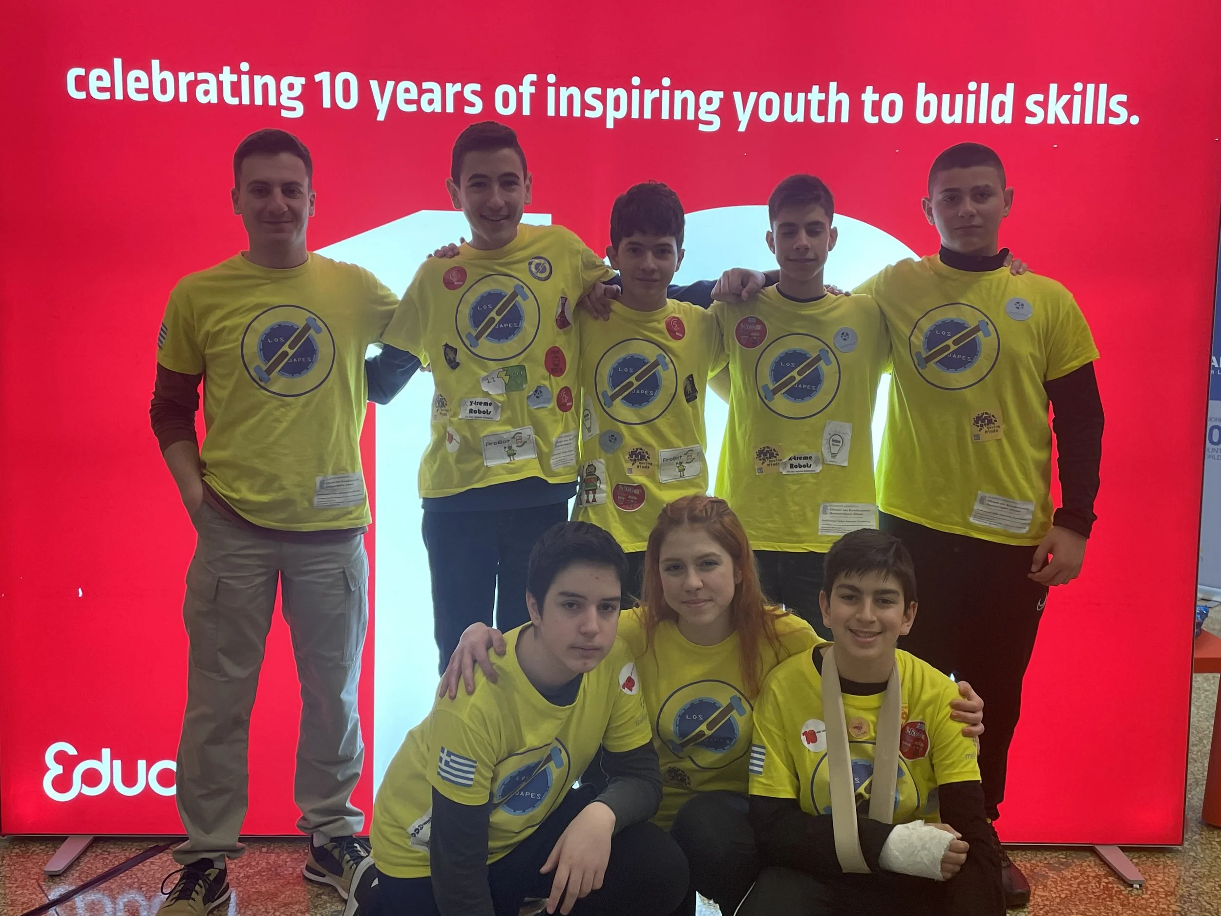 Group of five young people wearing yellow t-shirts with a logo, standing in front of a red background with text celebrating 10 years of inspiring youth to build skills, and posing for a photo.