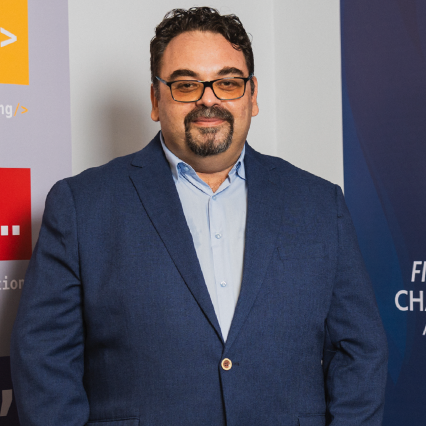 A man wearing glasses and a dark blue blazer smiling in front of a blue and gray background with signs related to coding, competition, and athletic challenges.