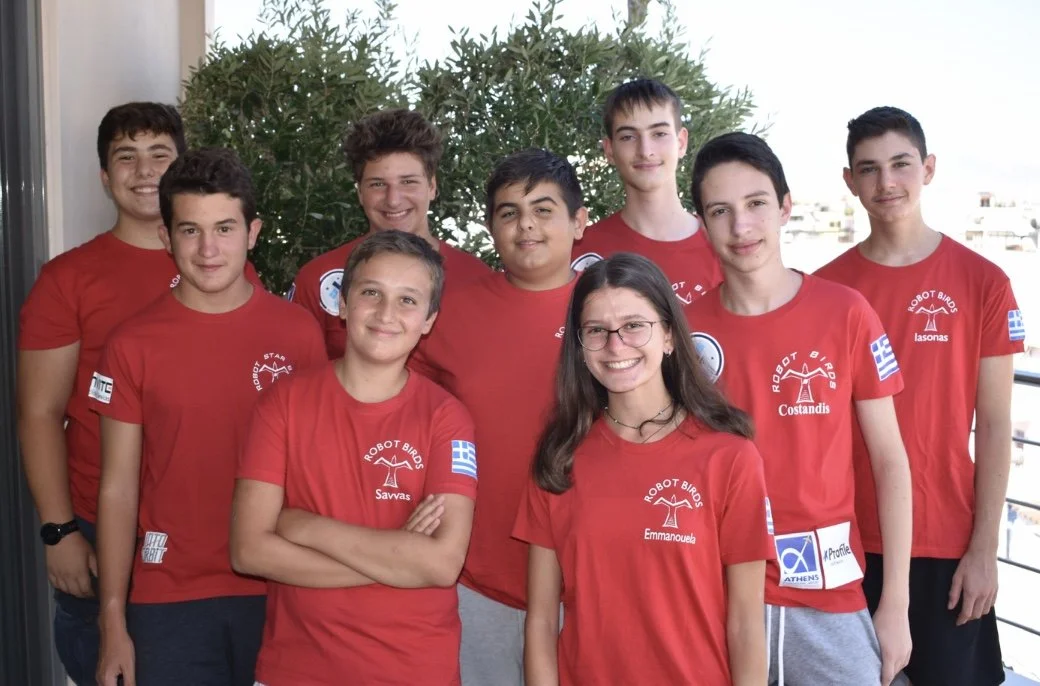 Group of nine young teenagers standing outdoors, wearing matching red T-shirts with a logo and Greek flag patch, smiling at the camera.