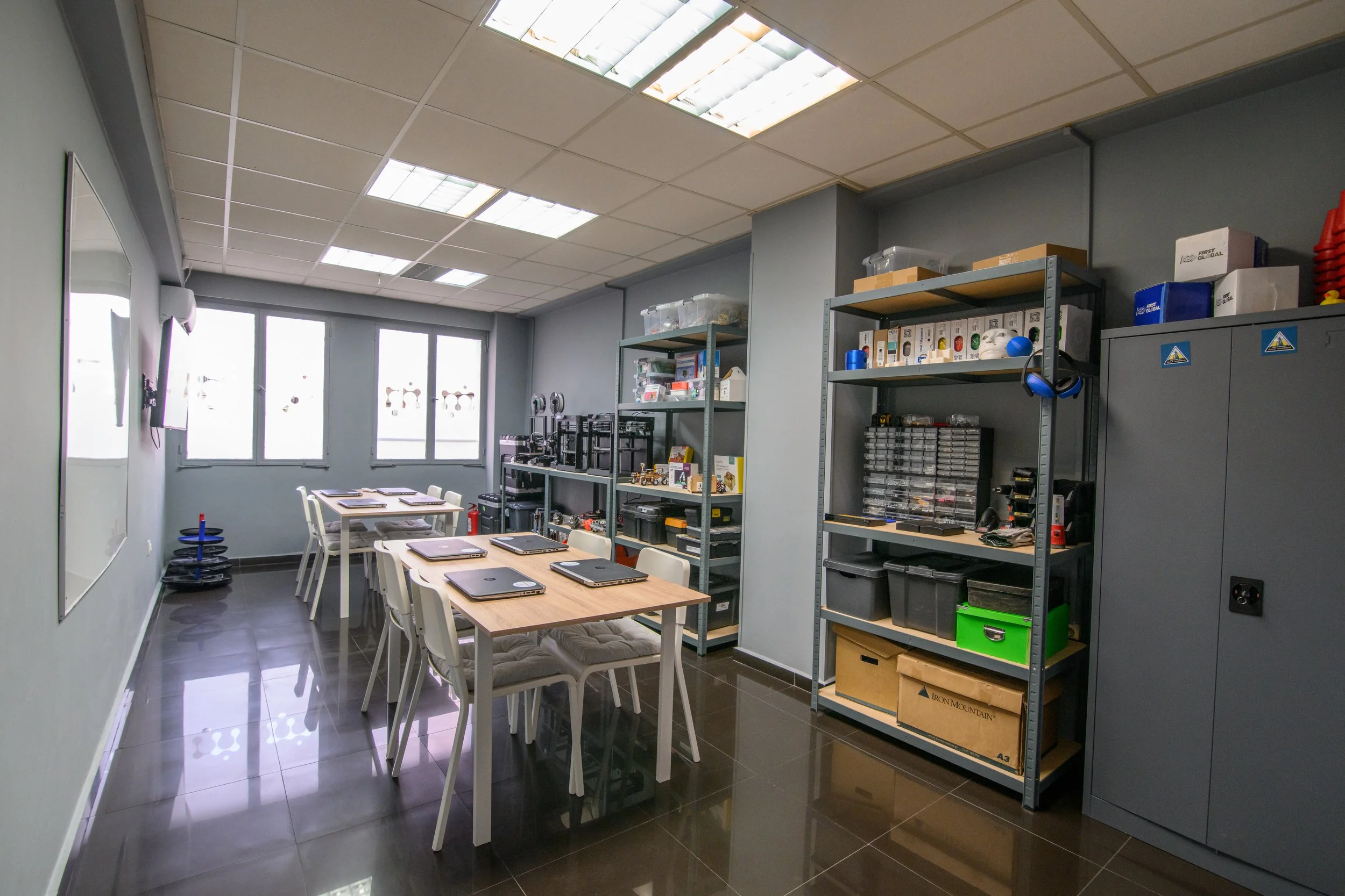 A conference room with a whiteboard, chairs around a table with laptops, and metal shelves filled with boxes and tools. The room has gray walls, a tiled ceiling with fluorescent lights, and windows letting in natural light.