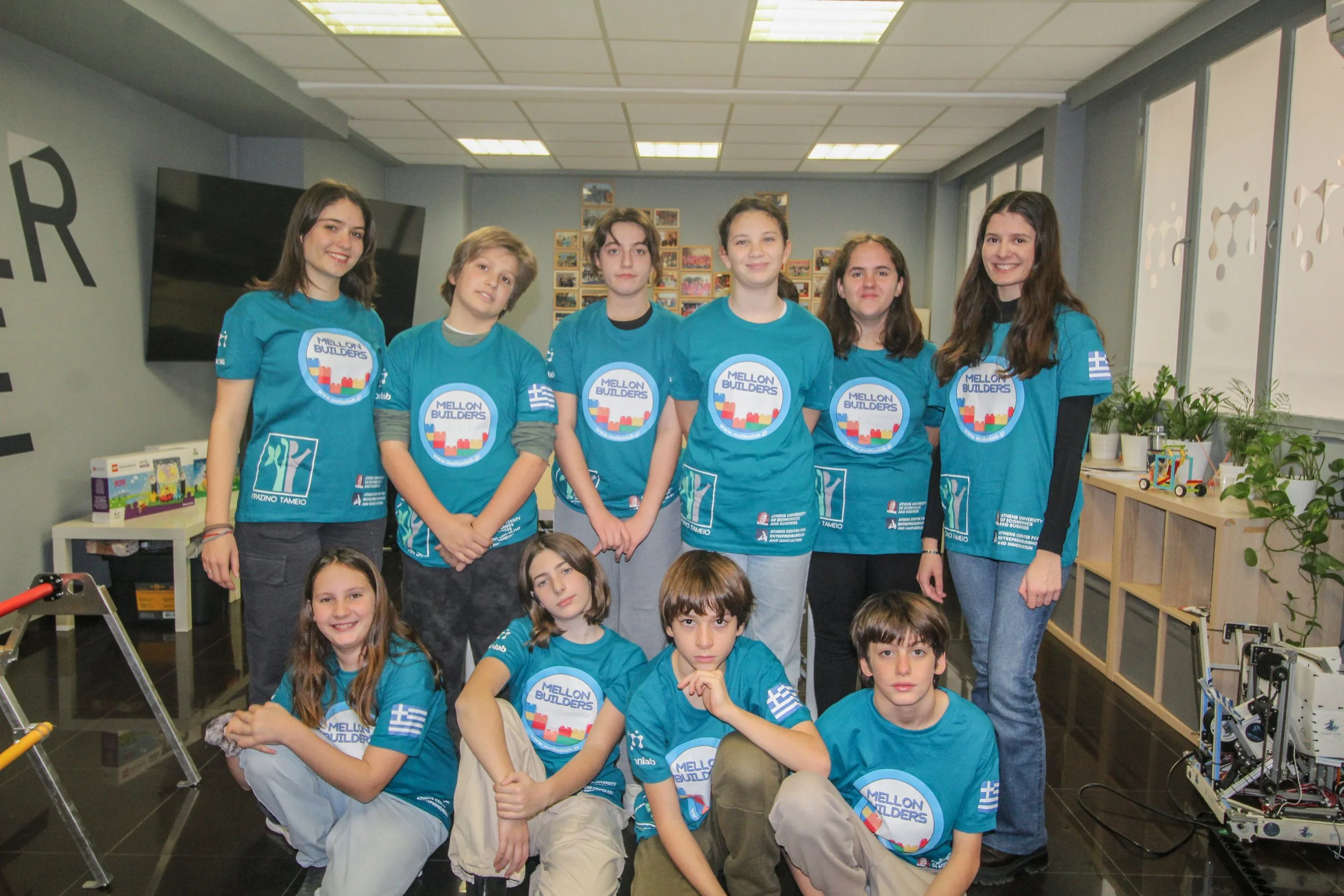 Group of young students in blue T-shirts standing together in a classroom or lab with a display of projects and photos in the background.