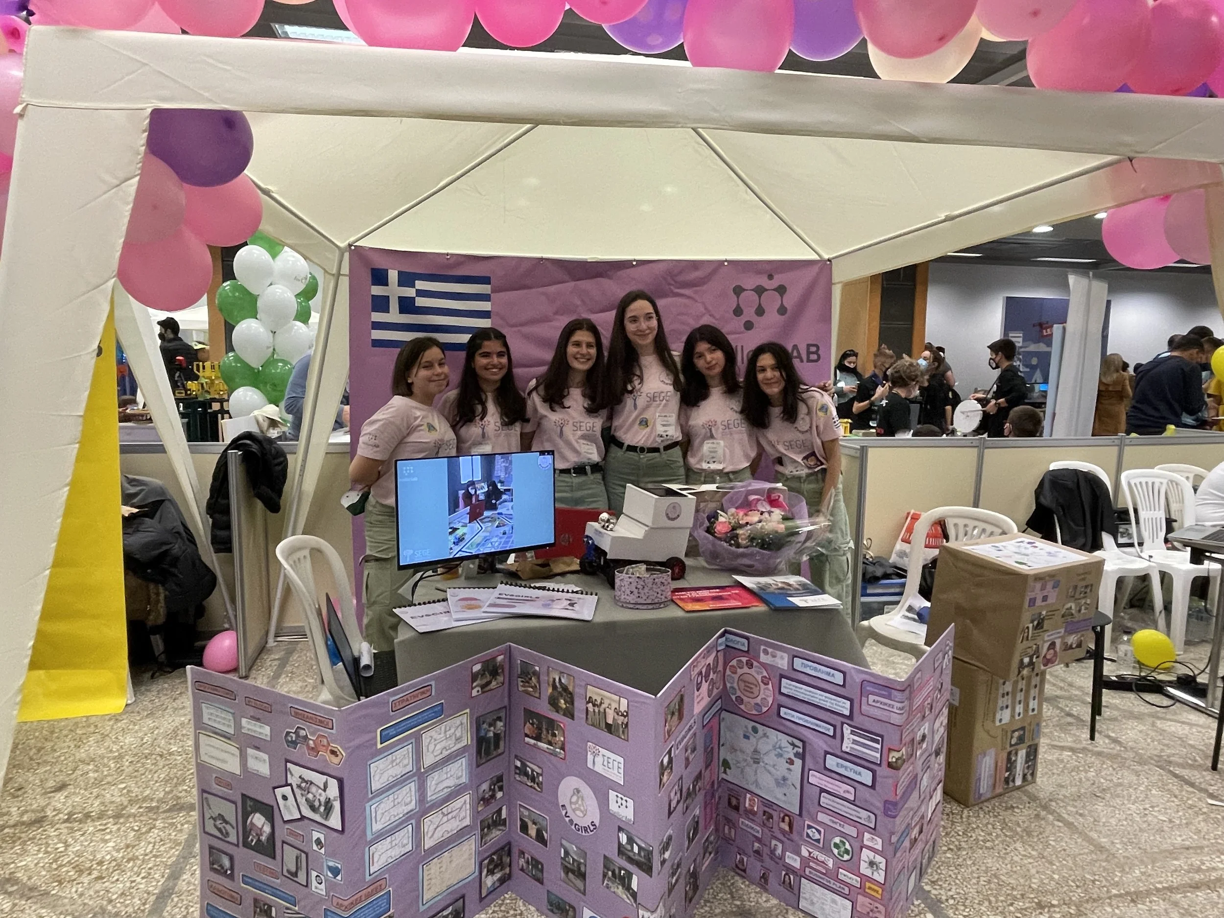 Five women standing in front of a purple display board at a booth with balloons and a Greek flag in the background. The women are smiling and wearing matching light pink shirts.