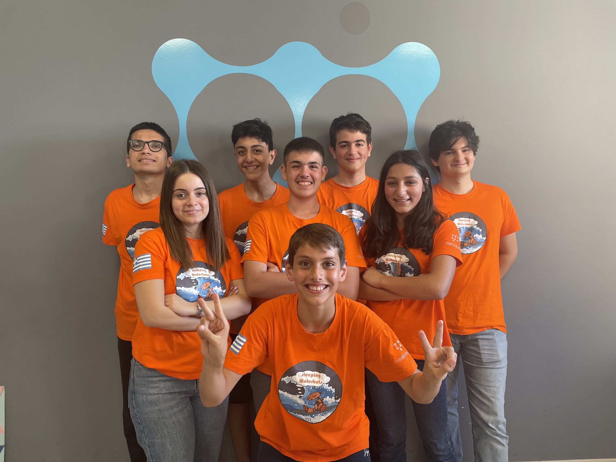 A group of seven diverse teenagers wearing orange shirts with a graphic of a cartoon waterdrop and the words 'Sleeping Waterboy' are standing in front of a gray wall with a blue abstract design. They are smiling and posing for a photo, with some making peace signs.