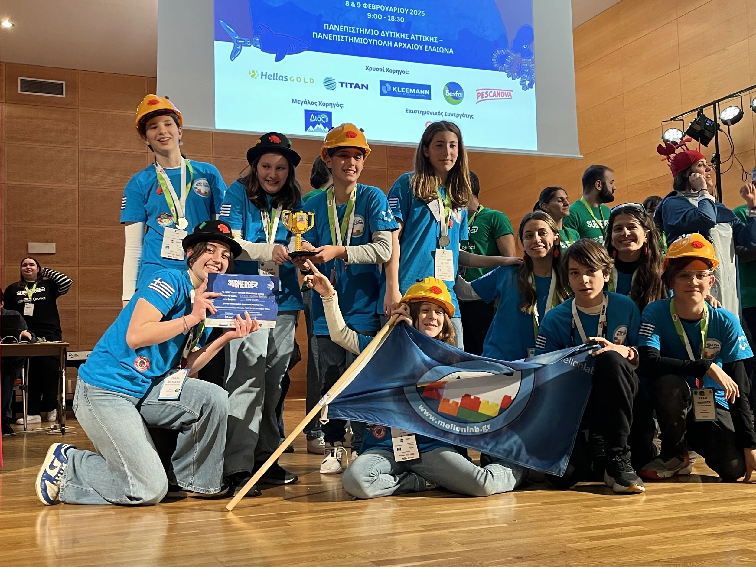 Children and young teens at an award ceremony, some wearing yellow helmets, some holding a flag and medals, celebrating on stage with a large screen in the background displaying event details in Greek and logos of sponsors.