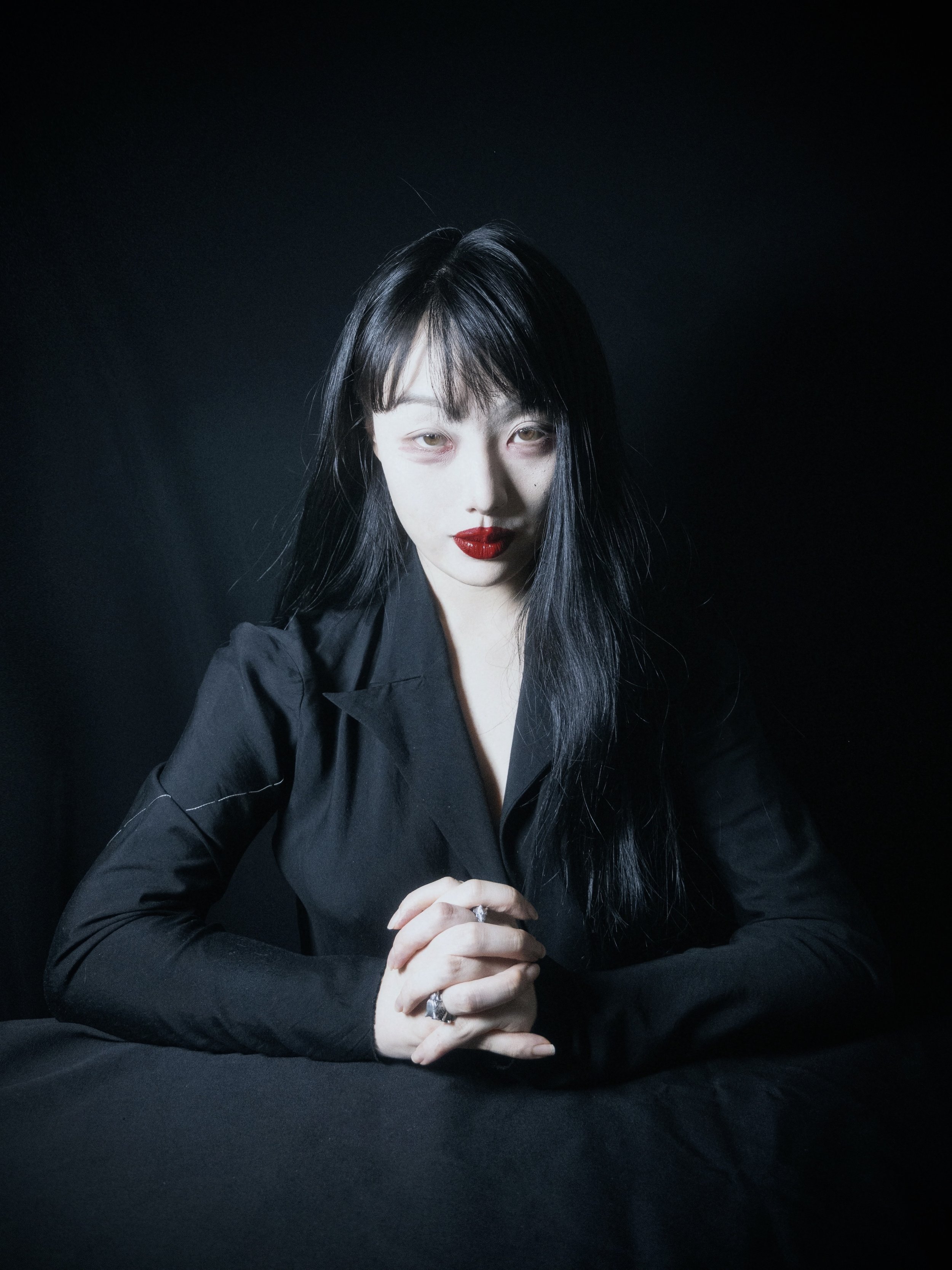 A woman with pale skin and long black hair, wearing red lipstick and rings, dressed in black, against a black background.