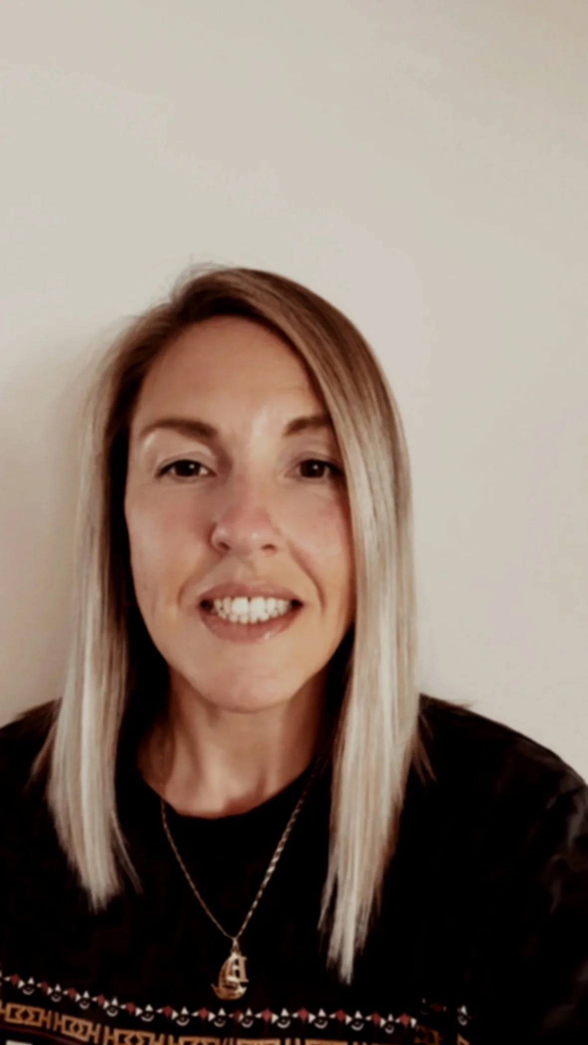 A woman with long straight blonde hair smiling, wearing a black top and a silver necklace with a pendant, standing against a light-colored wall.