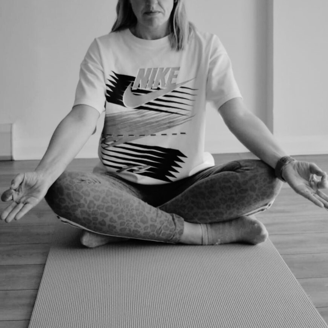 A woman sitting cross-legged on a yoga mat, practicing meditation with her hands resting on her knees, in a room with hardwood floors and plain walls.