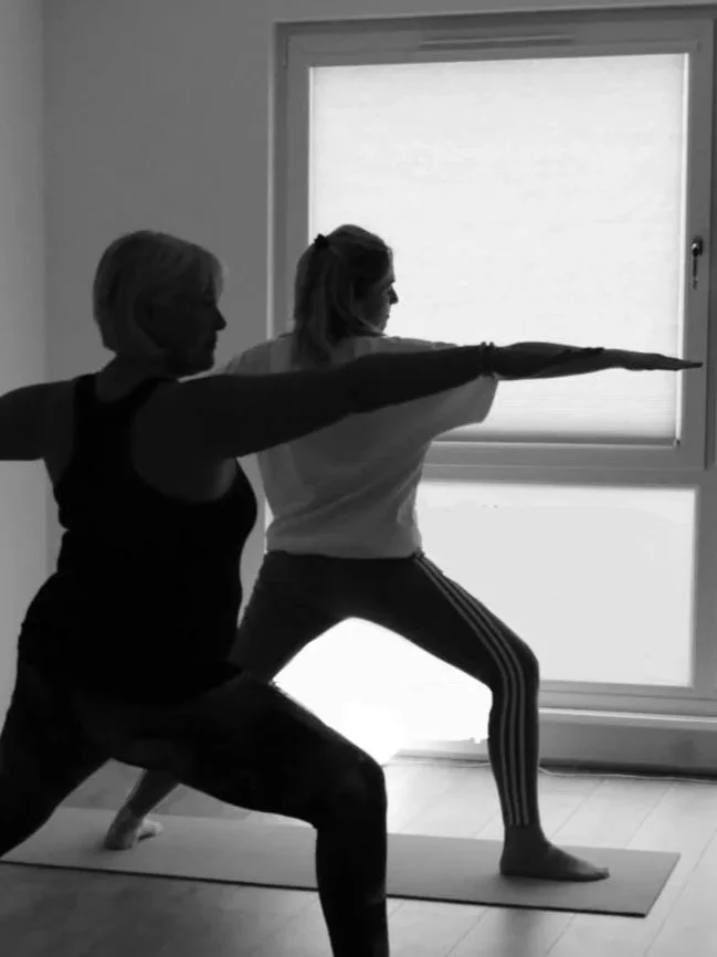 Two women practicing yoga indoors, one in a lunge pose with arms extended forward and the other assisting, near a large window.