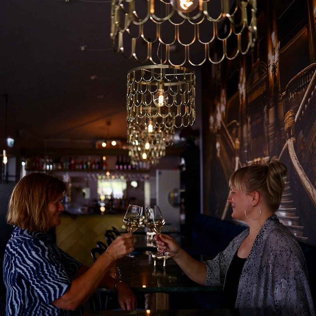 Två kvinnor som sitter vid ett barbord och skålar med glas vin i en bar eller restaurang med dämpad belysning och dekorativa lampor.