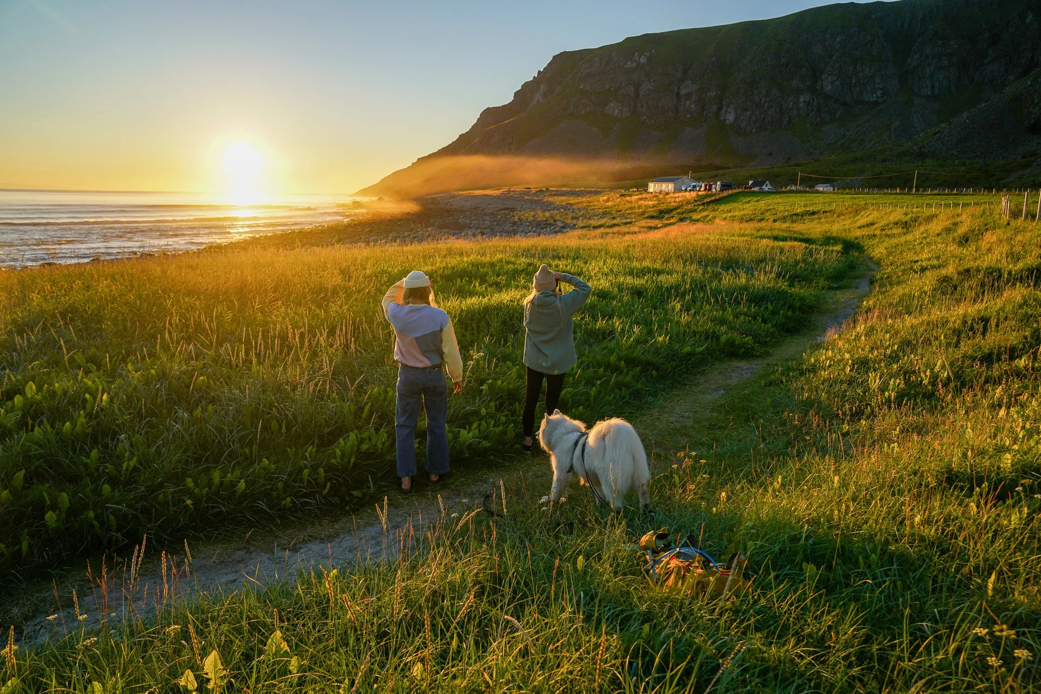 Two women and a dog walk along a beachside grassy trail during sunset, with the ocean on the left and mountains in the background.