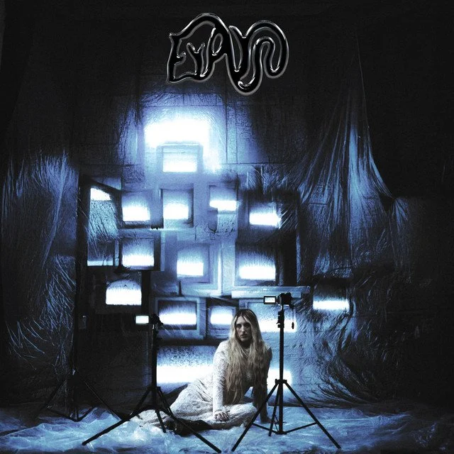 A woman with long blonde hair sitting on the floor in front of a camera setup, illuminated by multiple bright screens and a Zayn sign hanging above in a dark, artistic space.