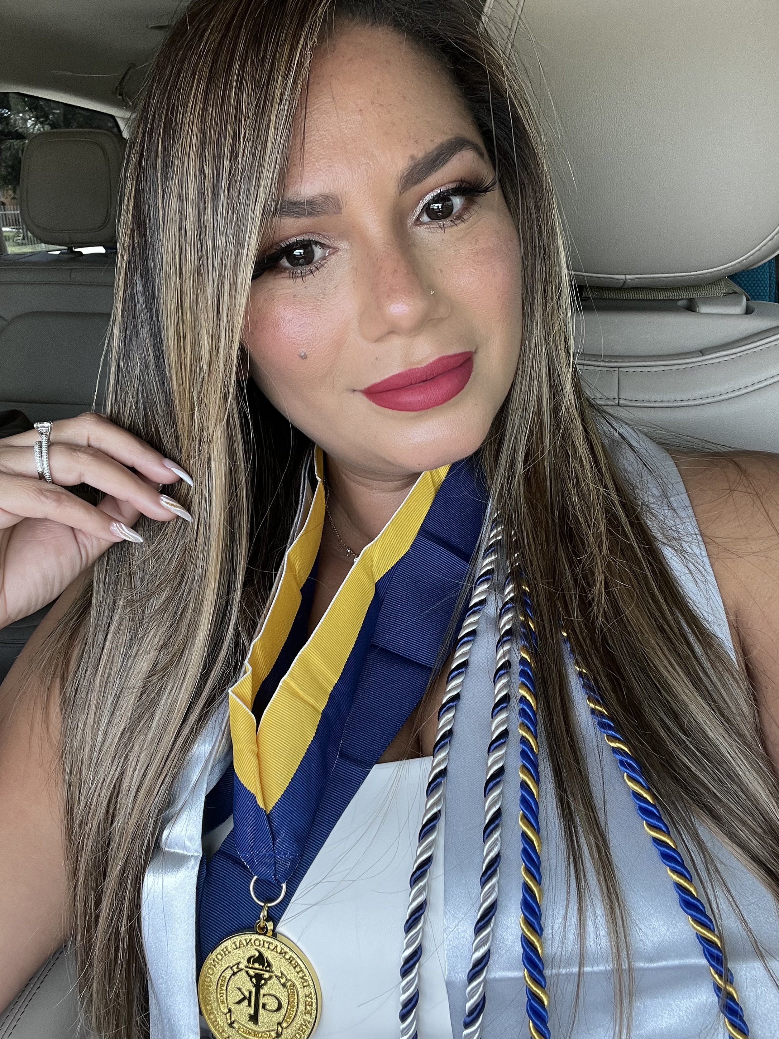 A woman with long, light brown hair and makeup, wearing a graduation stole with cords, a medal, and a ring, sitting inside a car.