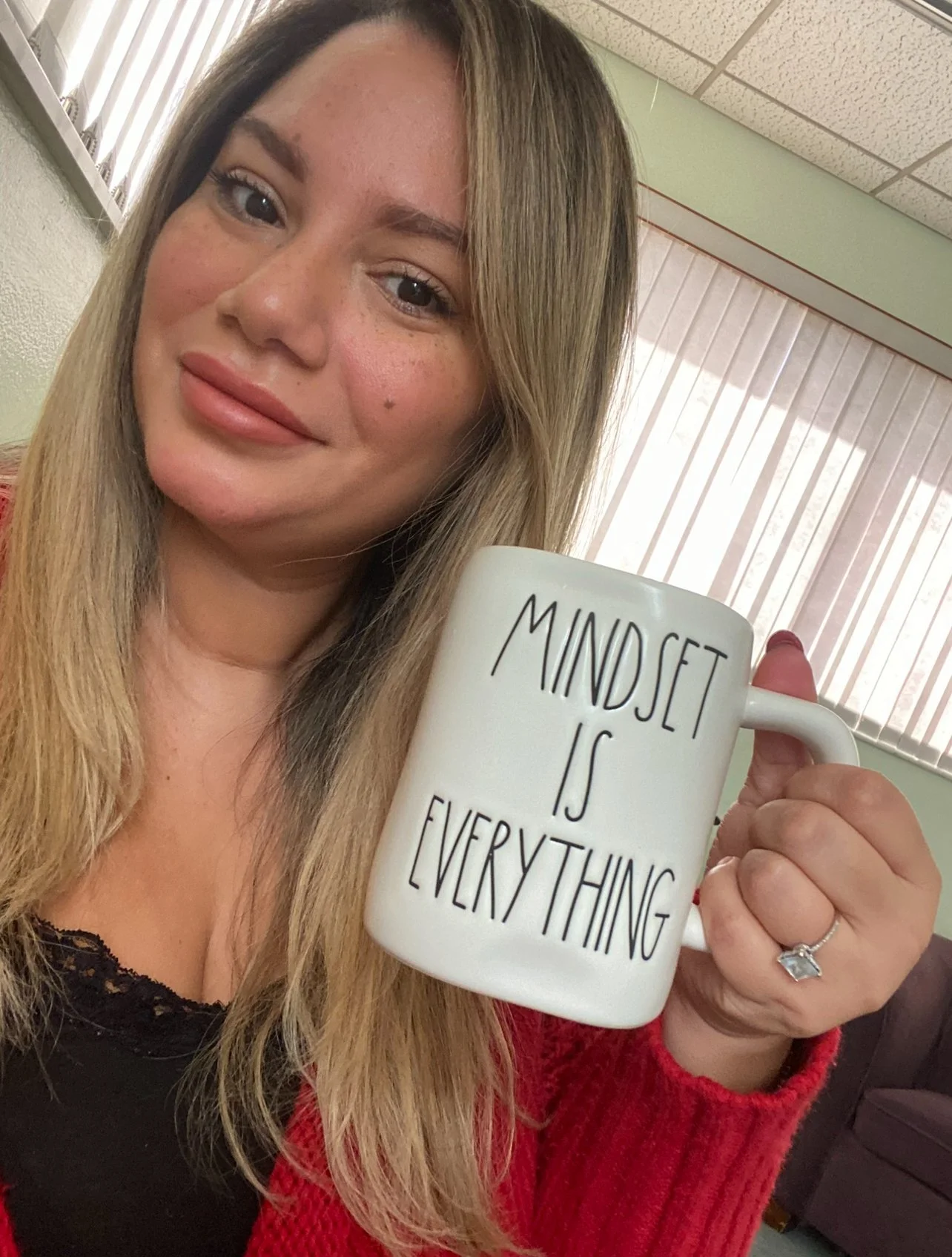 A woman with long blonde hair holding a white mug that says 'Mindset is Everything'. She has a warm inviting smile and she is wearing a red sweater.
