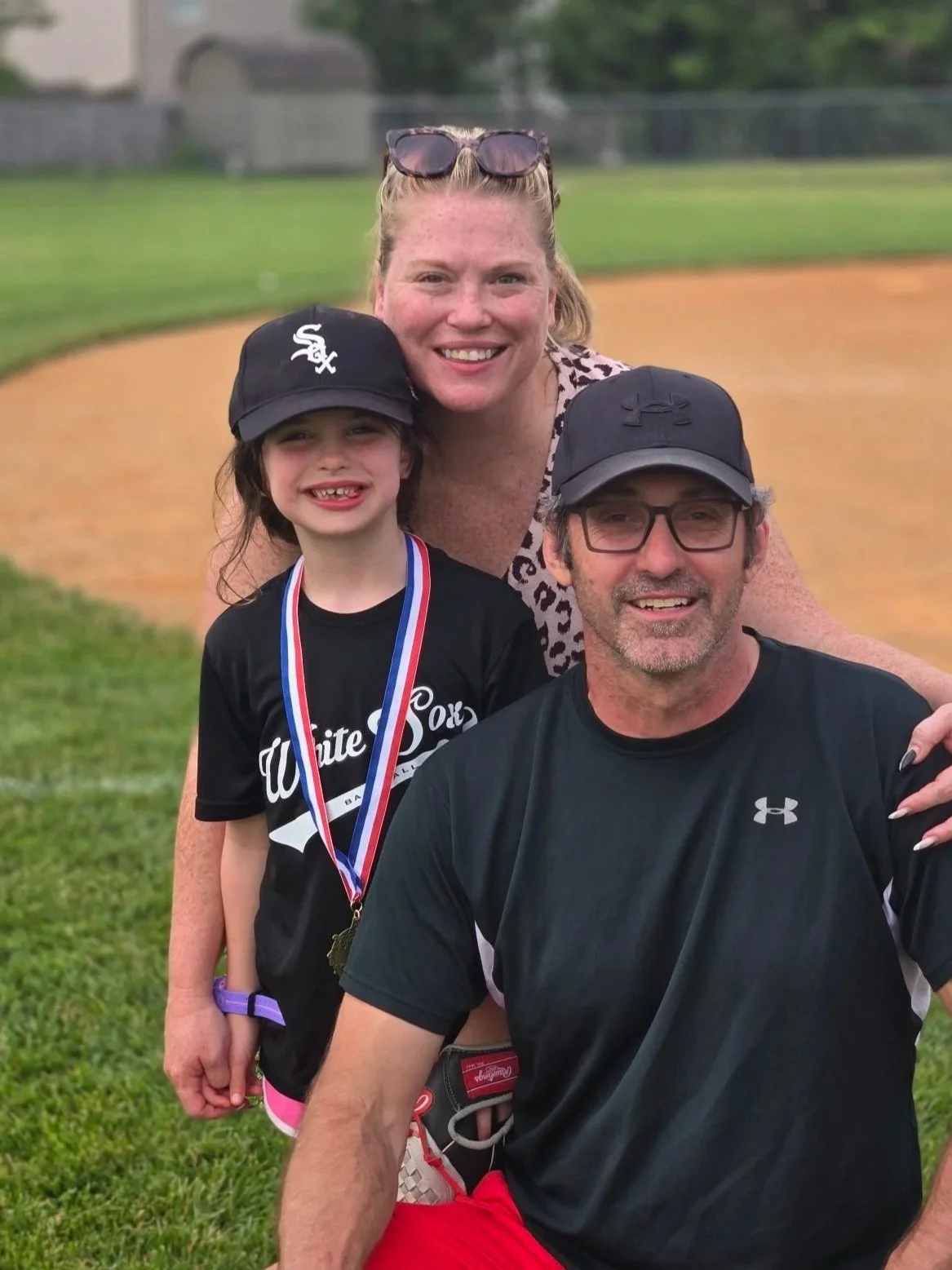 A family of three posing on a baseball field. The young girl has a medal around her neck, and they are smiling happily.