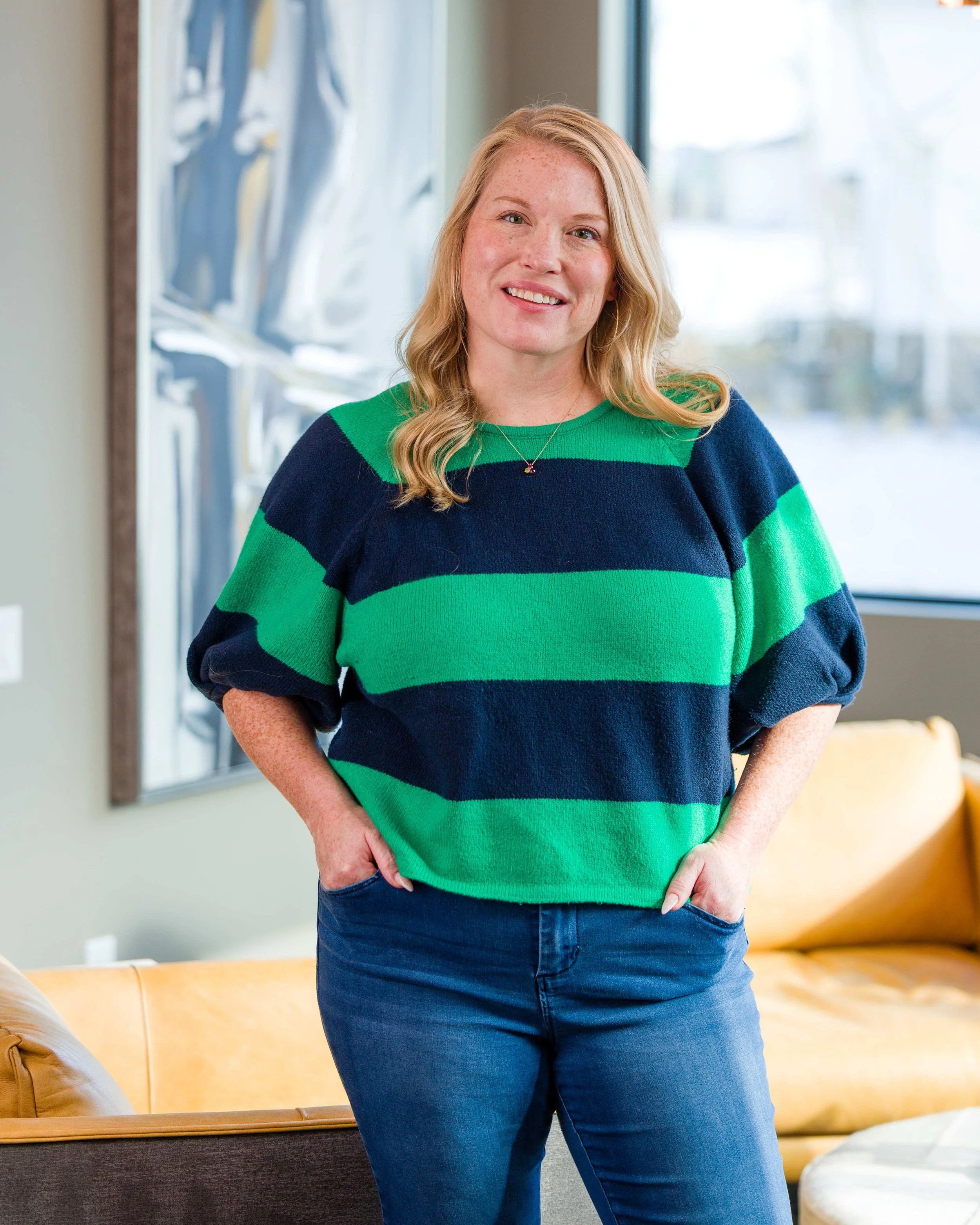 Stephanie Jo Yocum, wearing a green and navy blue striped sweater and blue jeans, standing indoors in front of a window with a view outside.