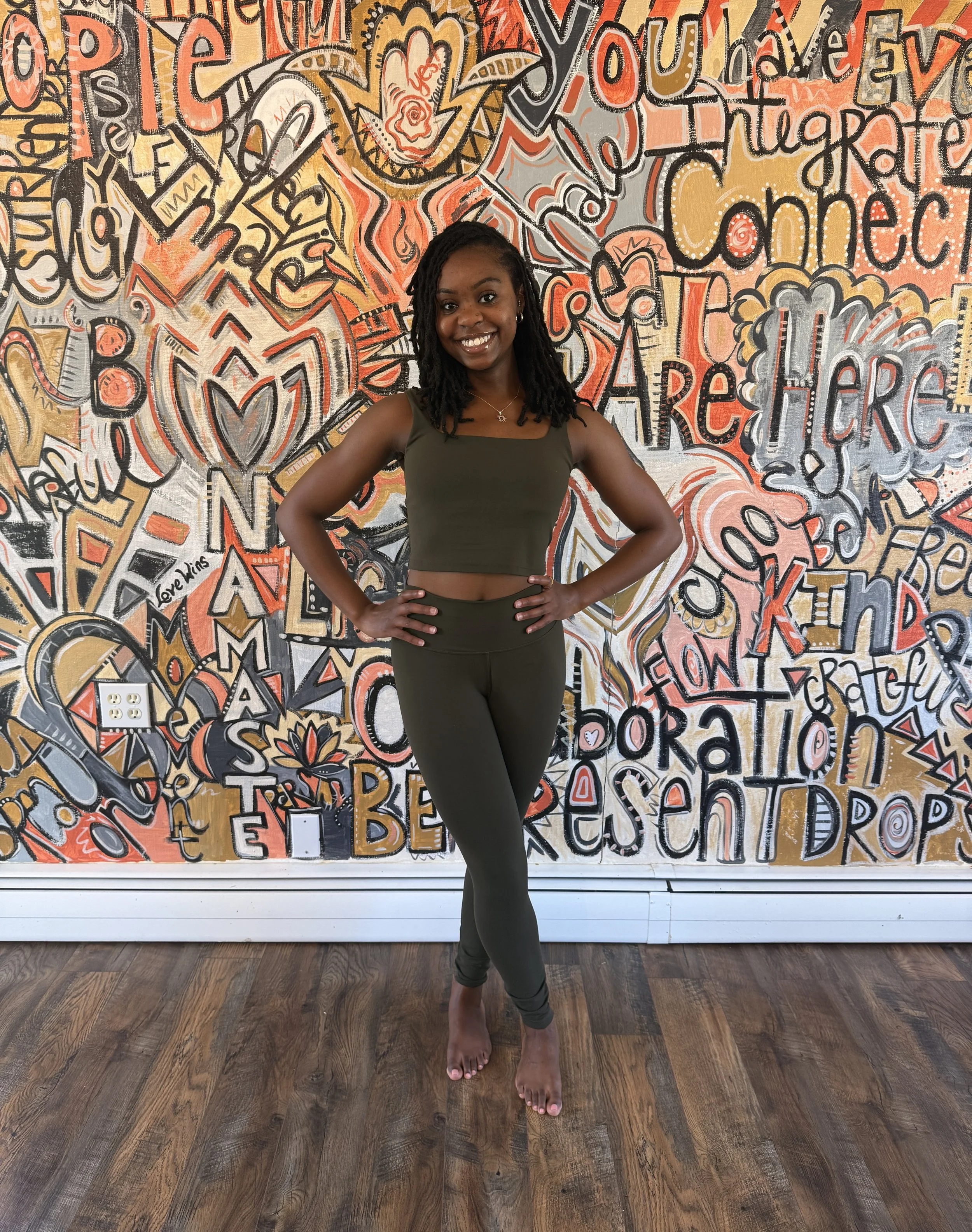 A woman with long hair smiling in front of a colorful graffiti mural with words like 'You Are Here,' 'Light,' and 'Create.'