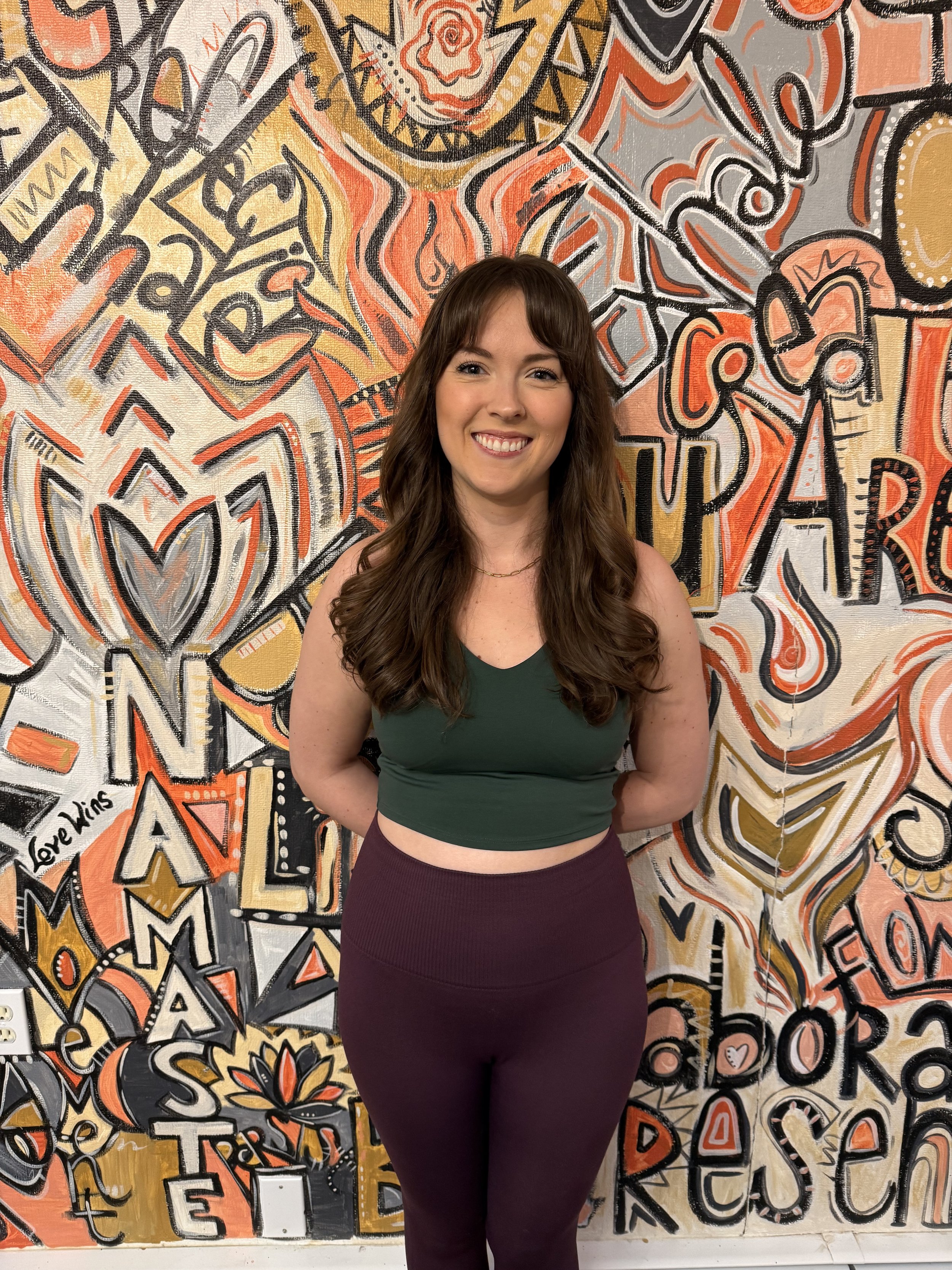 A smiling woman with long brown hair standing in front of a colorful abstract wall mural.