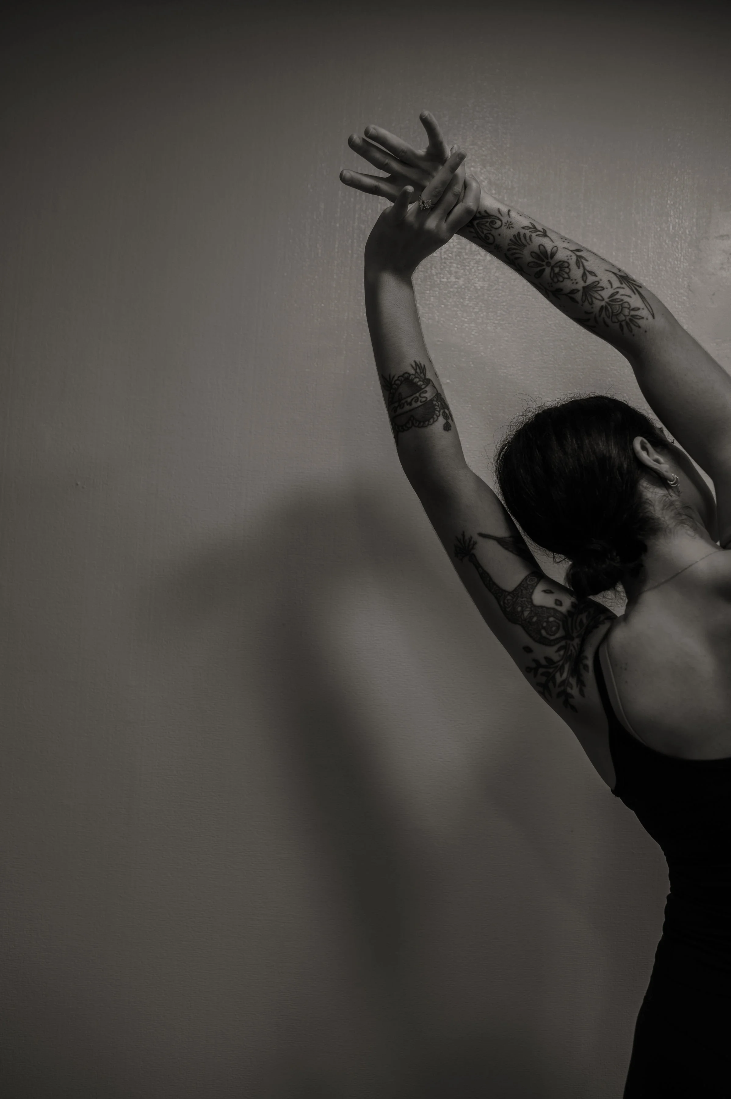 A woman with tattoos poses with her arms raised above her head in a black and white photo.