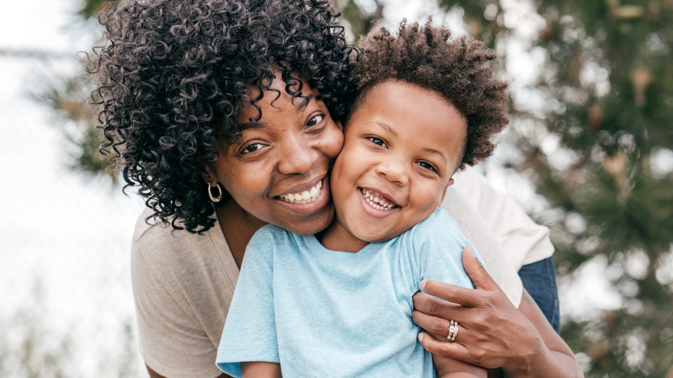 A joyful woman hugging a smiling young boy outdoors with trees in the background.