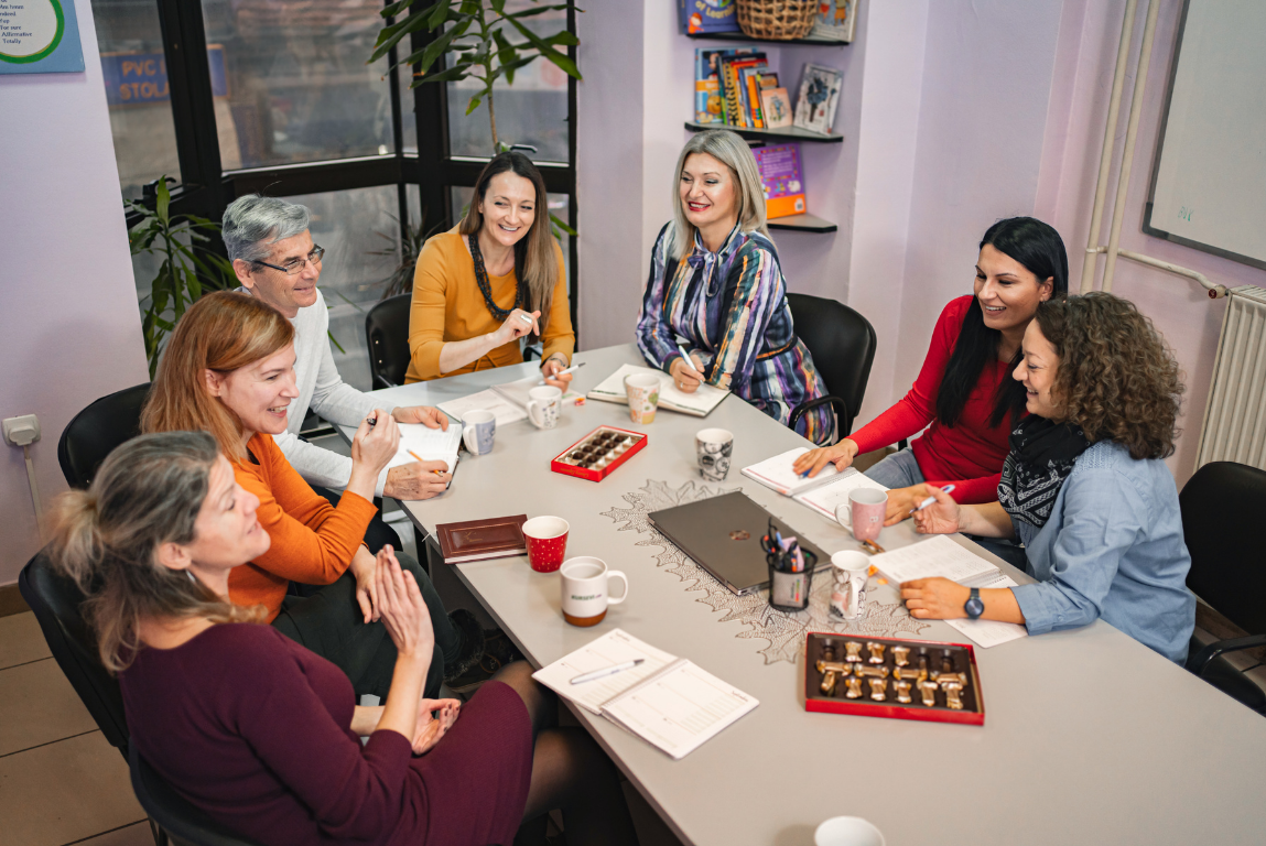 A diverse group of seven women sitting around a conference table, smiling and engaging in a discussion during a meeting.