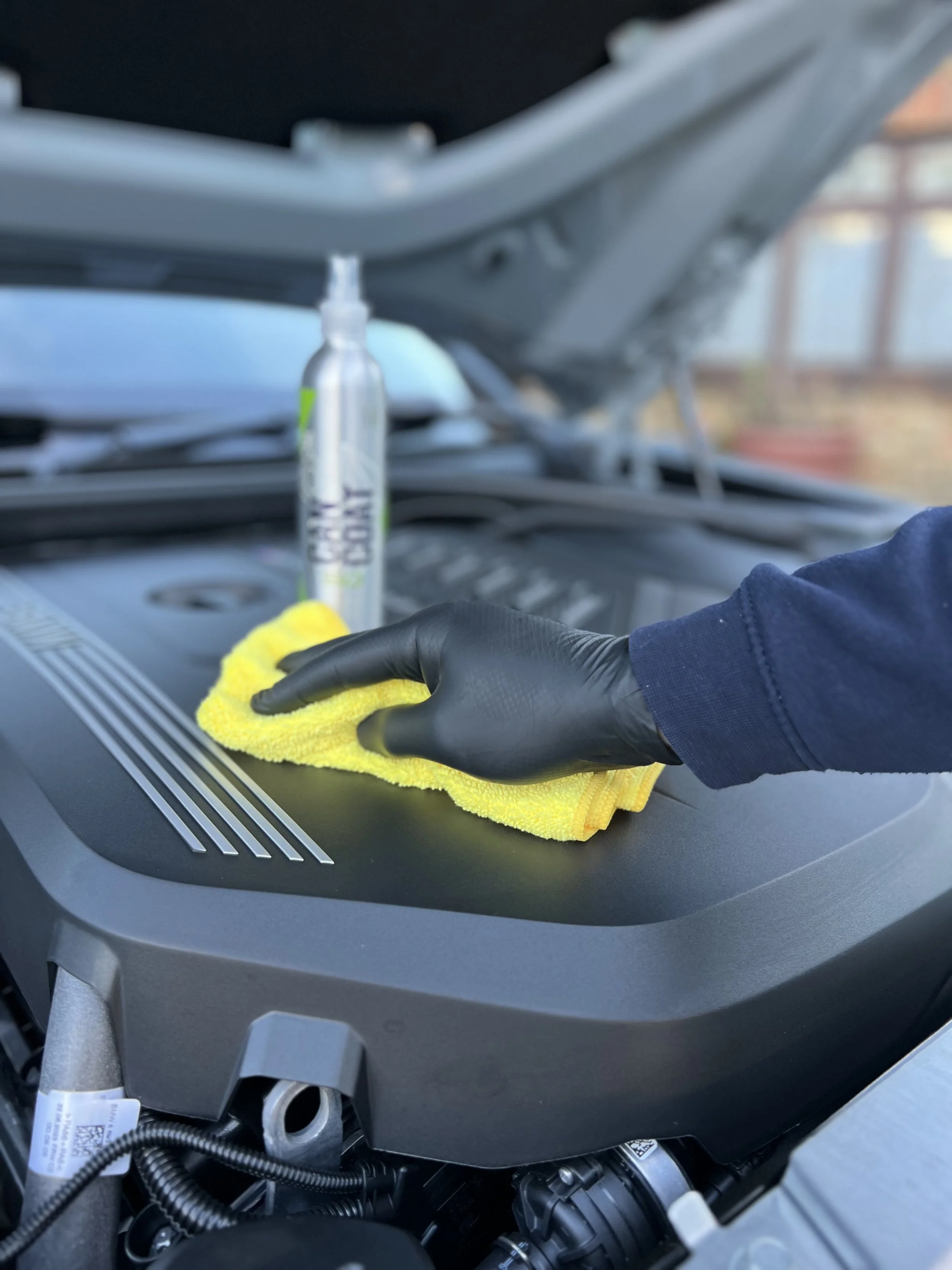 Person wearing a black glove wiping a car engine with a yellow cloth, with a cleaning spray bottle in the background.