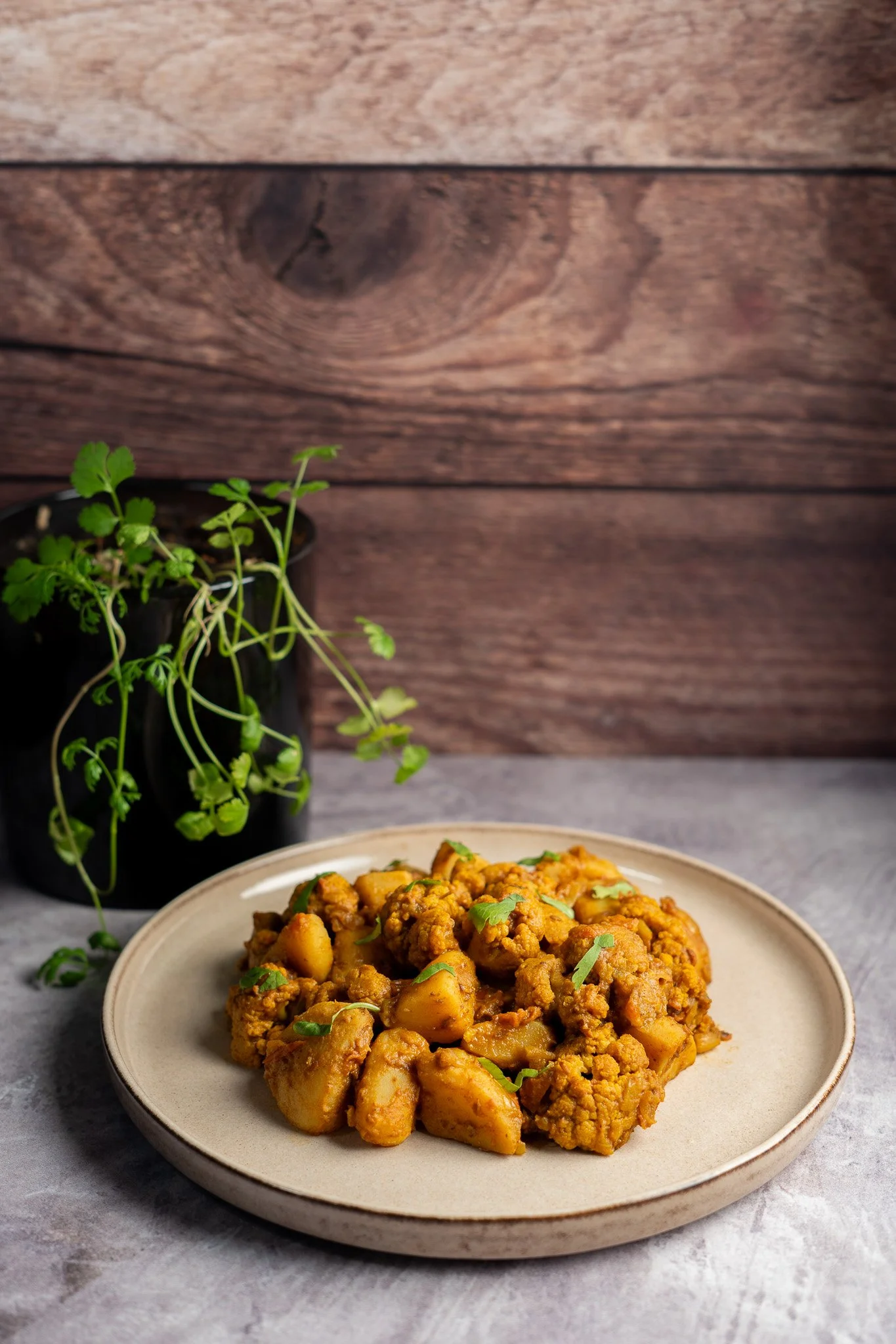 Plato de curry con papas y carne, decorado con cilantro, en un plato beige, con planta de hierbas al fondo sobre mesa gris y fondo de madera.