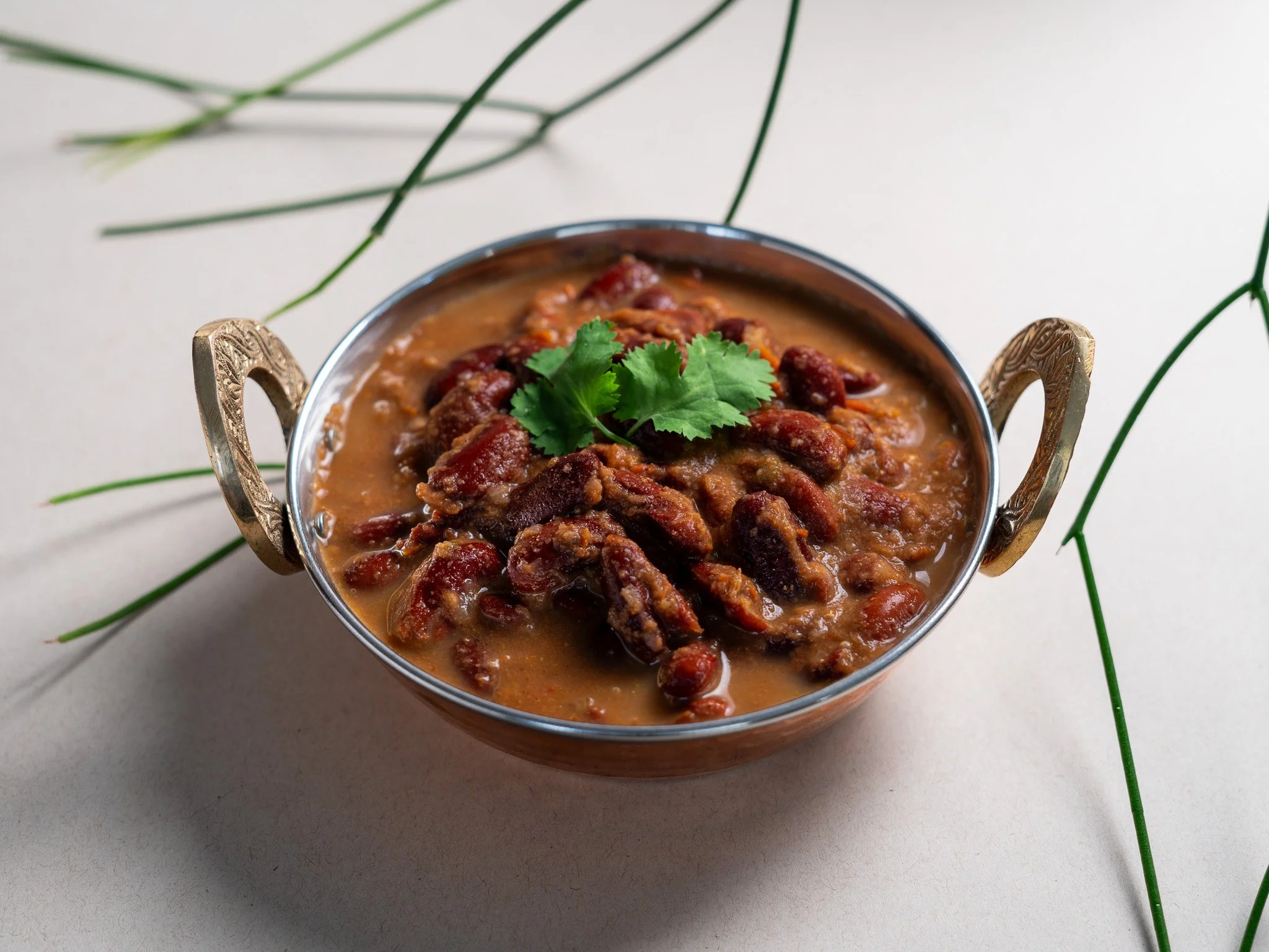 Plato de carne en salsa, decorado con cilantro, en un recipiente de metal con asas doradas, sobre fondo claro con hojas verdes.