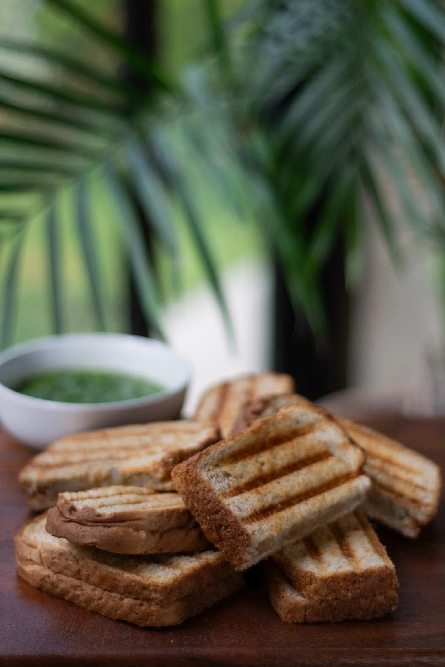 Tostadas con mantequilla y un plato con salsa verde, sobre una mesa de madera, y plantas verdes en el fondo.