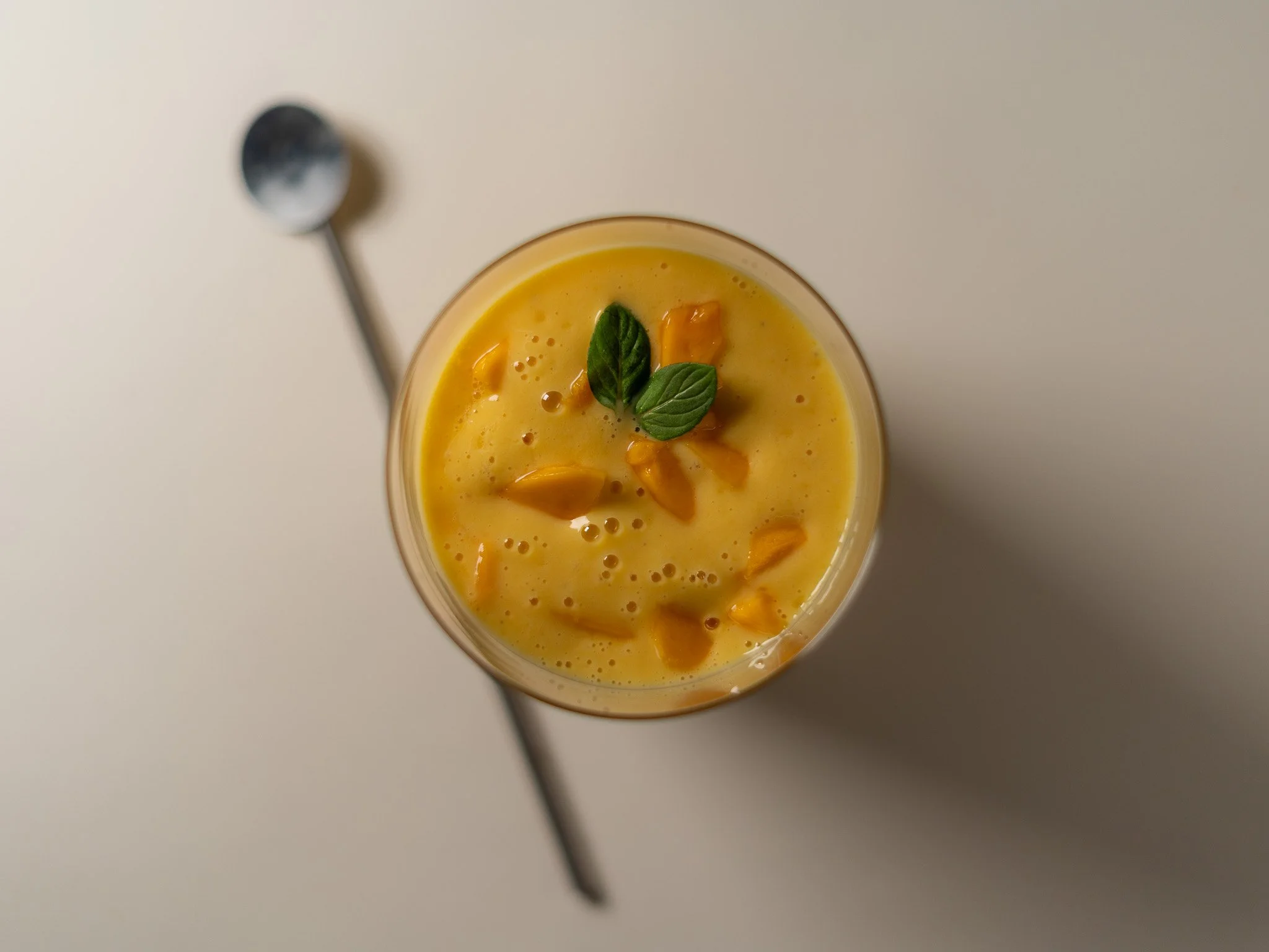 Vaso con bebida amarilla decorada con hojas de menta y trozos de fruta, sobre mesa blanca con cuchara de metal al lado.