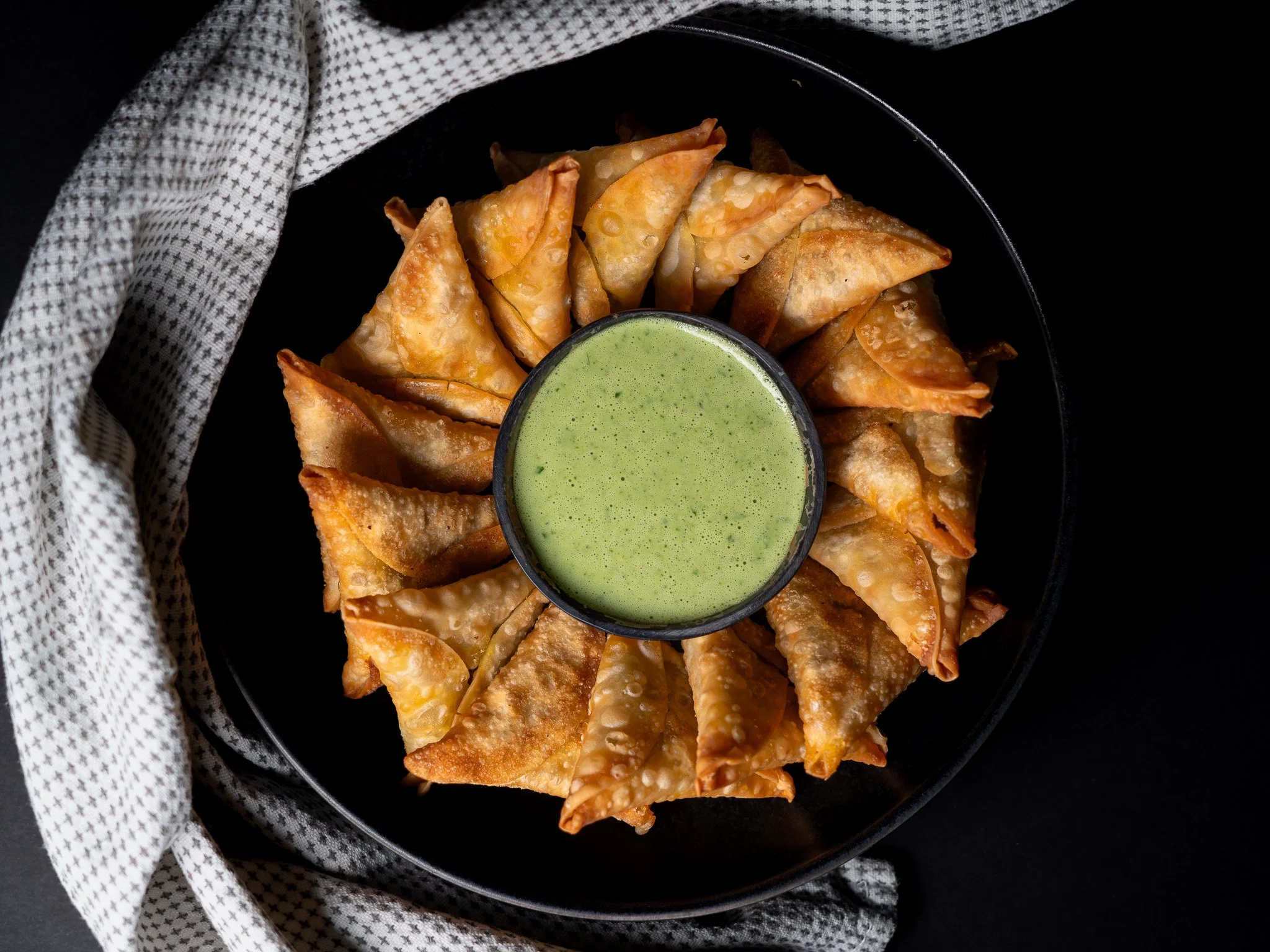 Plato de empanaditas fritas en forma de triángulo con salsa verde en recipiente central, sobre plato negro con mantel de tela a cuadros blanco y negro.